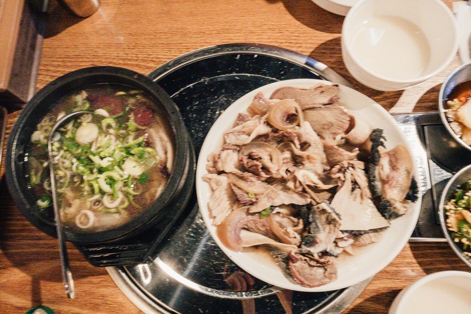 Overhead view of a mixed platter of boiled meats next to a bubbling stone pot of hangover soup, garnished with sliced scallions