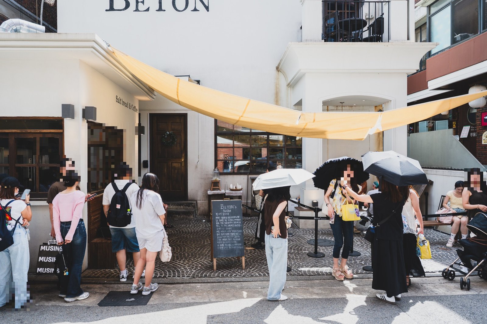 The dine-in queue outside BETON on a sunny weekend afternoon, several customers holding umbrellas for shade while waiting