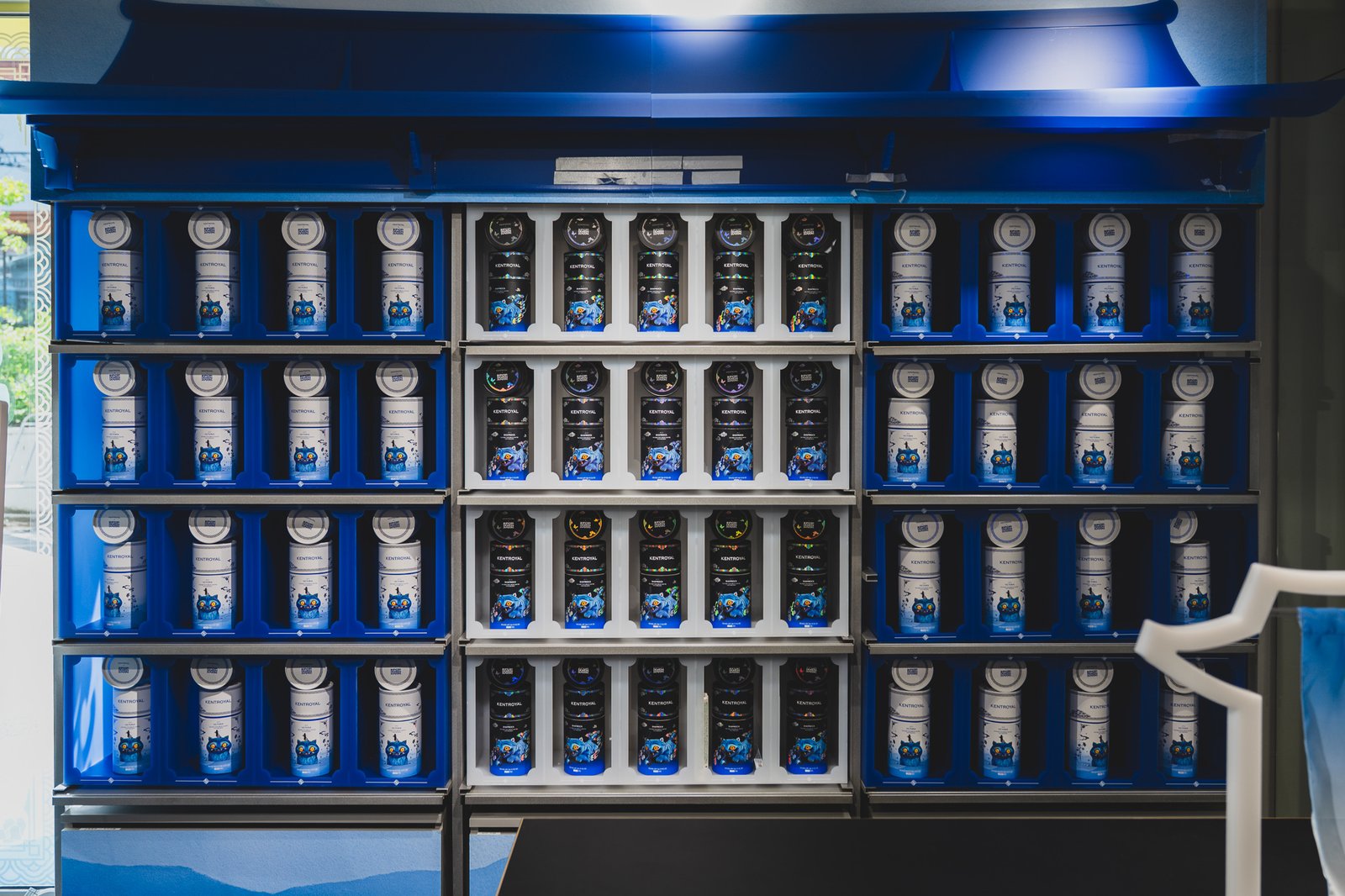 A tiered display wall with holographic collab tins in the center black section and standard-line tins symmetrically arranged in the white and blue sections on either side