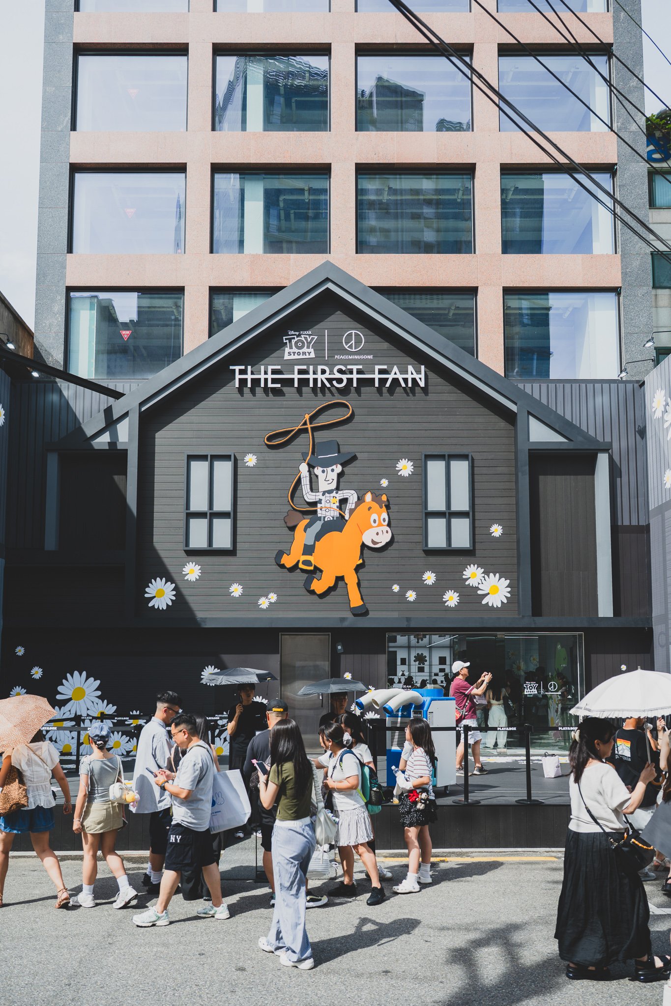 Exterior of THE FIRST FAN pop-up, a house-shaped facade with a "THE FIRST FAN" sign, a large Woody-on-horse graphic surrounded by daisies, and a long queue of visitors holding umbrellas against the sun