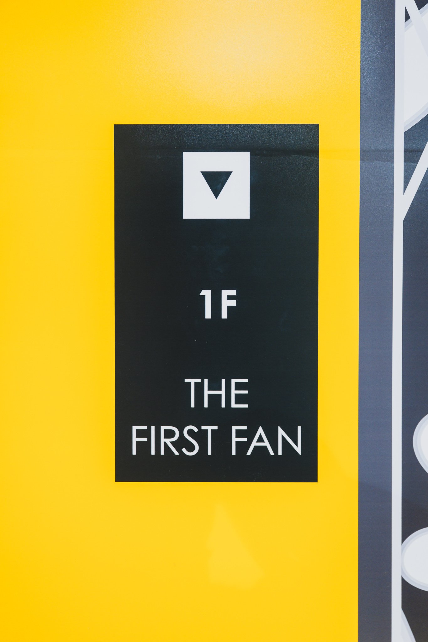 Black-and-yellow floor signage reading "1F THE FIRST FAN" with a down arrow, marking the transition from the 2nd floor shopping area to the 1st floor experience space