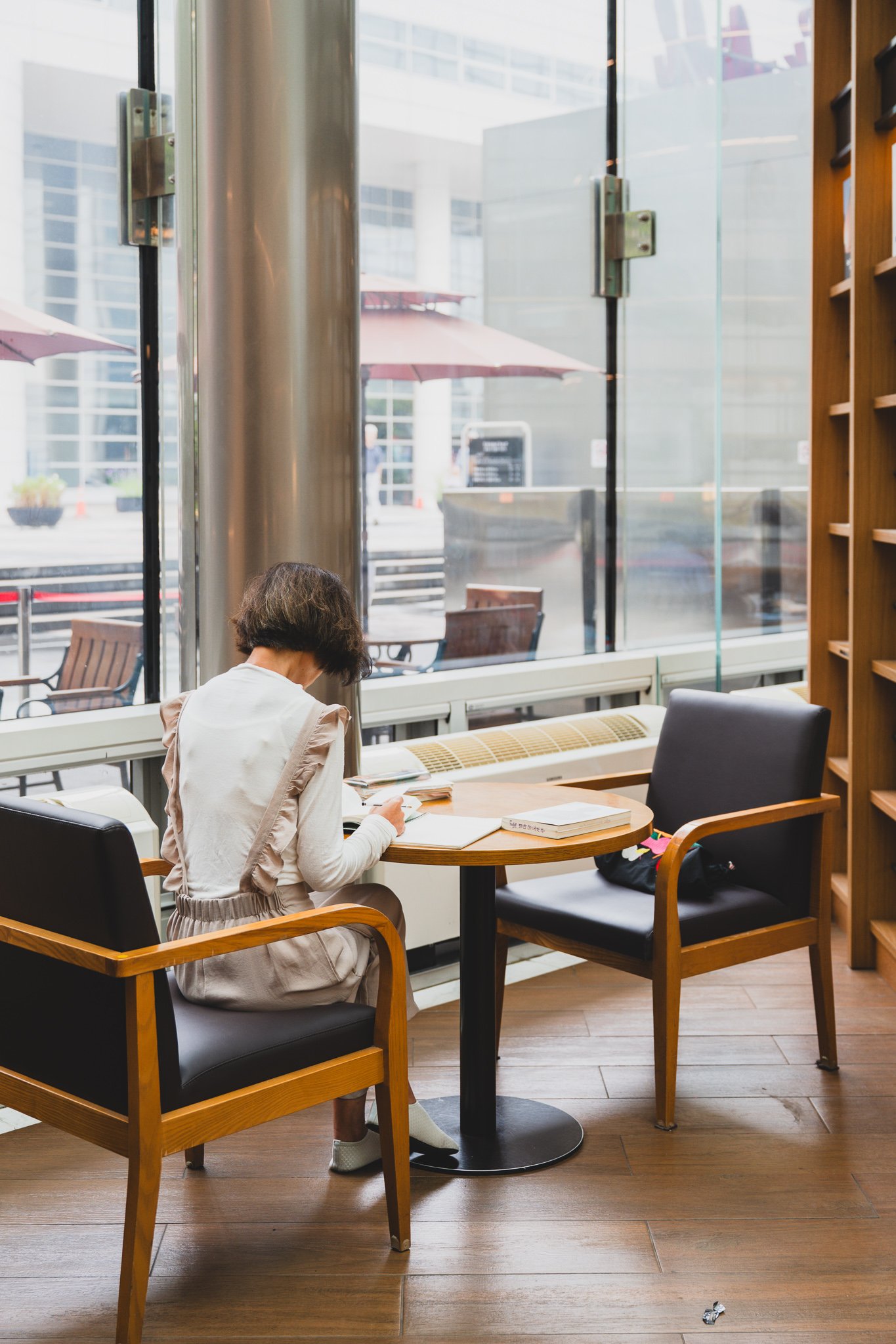 A woman reading alone at a small window-side table, with cafe chairs tucked into a quiet corner away from the main plaza
