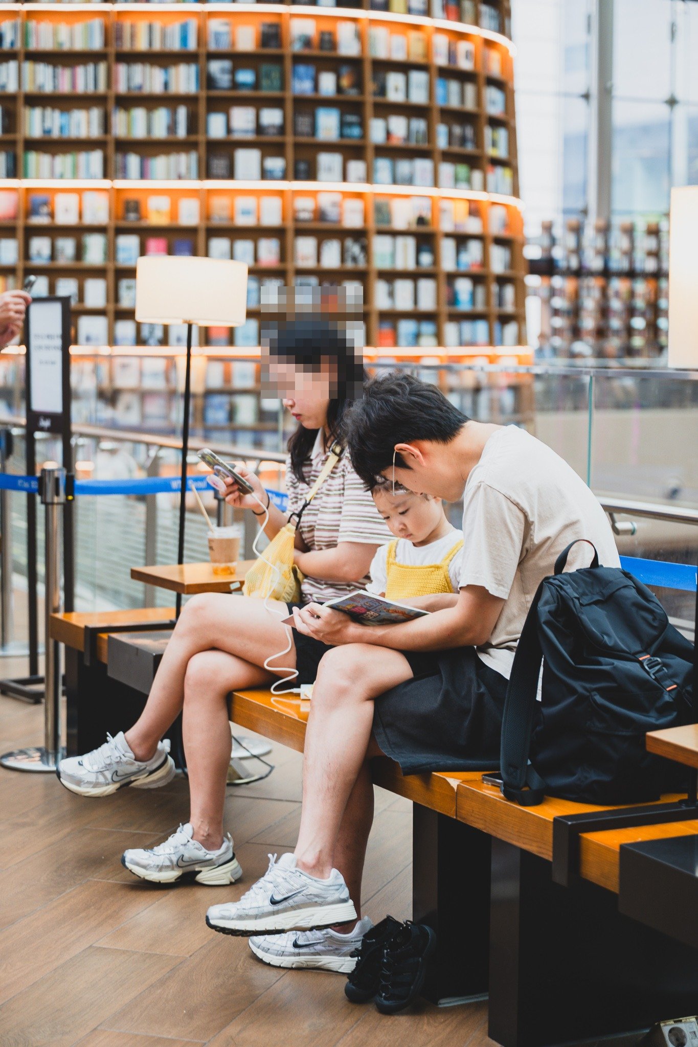 A family sharing a bench seat by the windows, an adult and child reading together with a backpack resting beside them