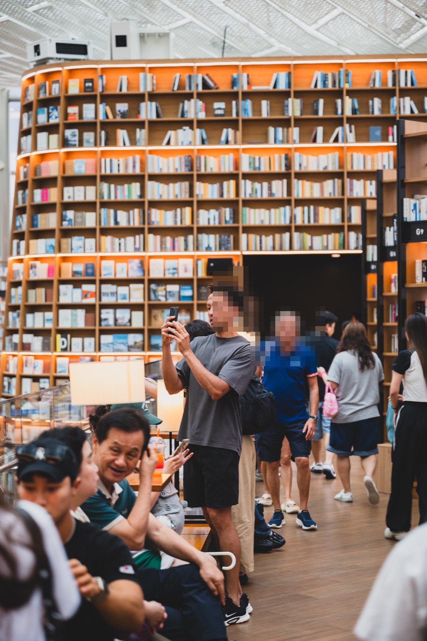 A visitor photographing the bookshelf tower on his phone, with other tourists and families milling around the base of the shelves