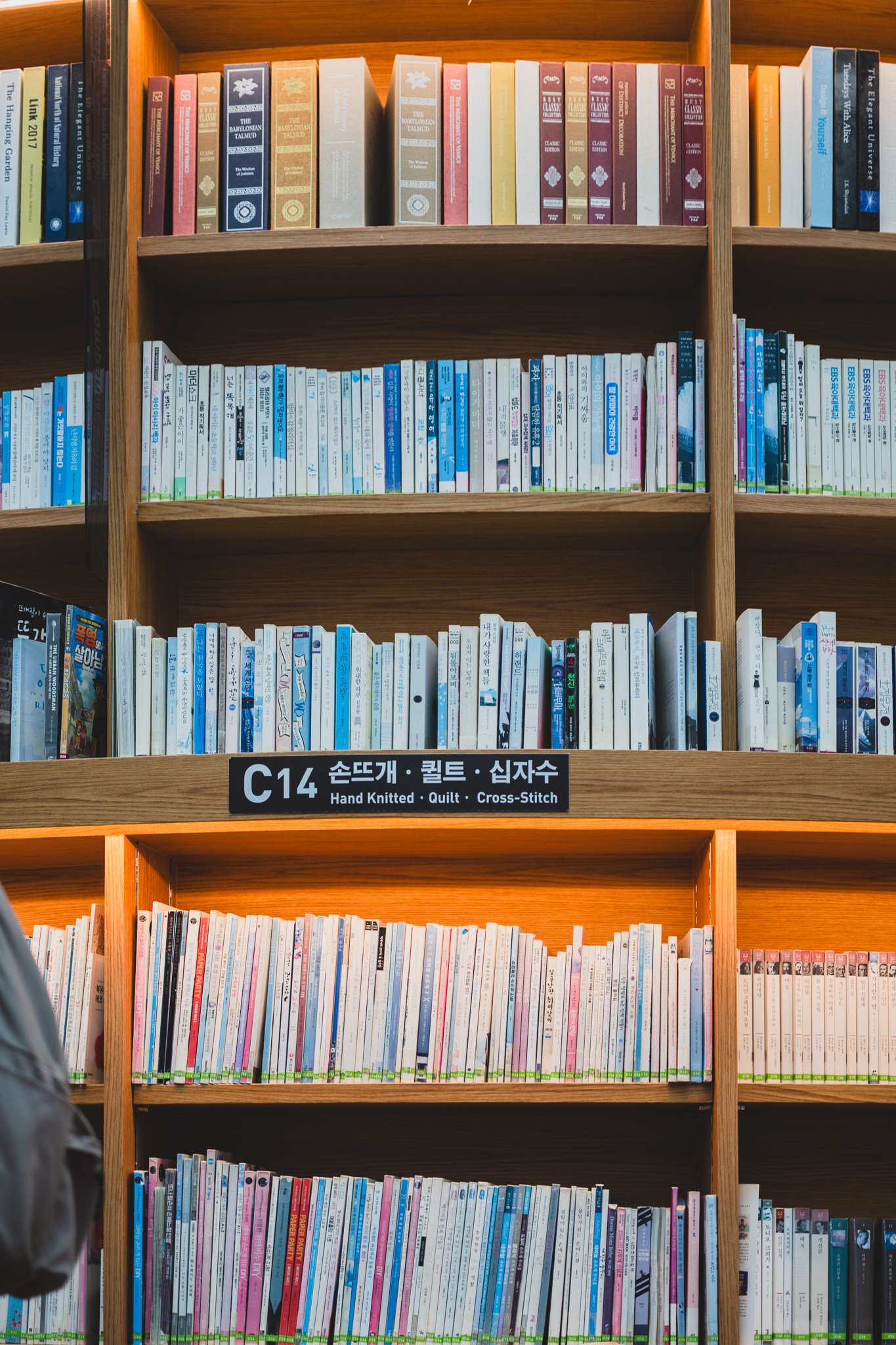 A close-up bilingual shelf sign reading "C14 Hand Knitted · Quilt · Cross-Stitch," with a dedicated hobby-book section underneath it
