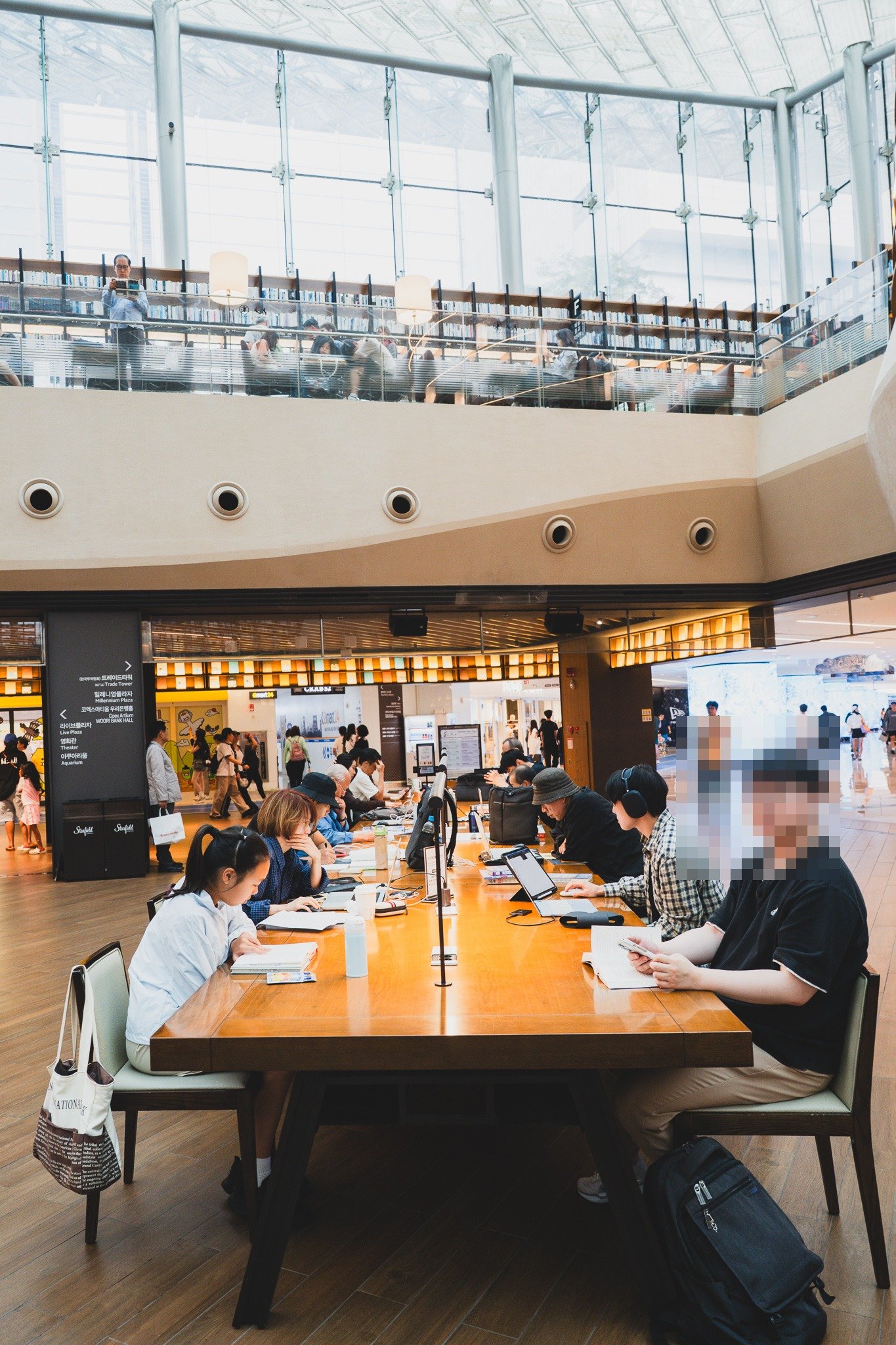A long communal table where several people sit working on laptops and tablets, with bags and drinks on the table between them