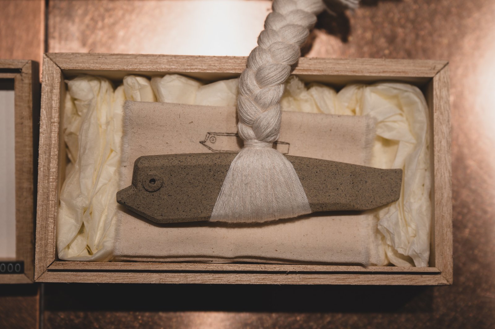 A small ceramic charm packaged in a wooden gift box lined with tissue paper and tied with a braided cord
