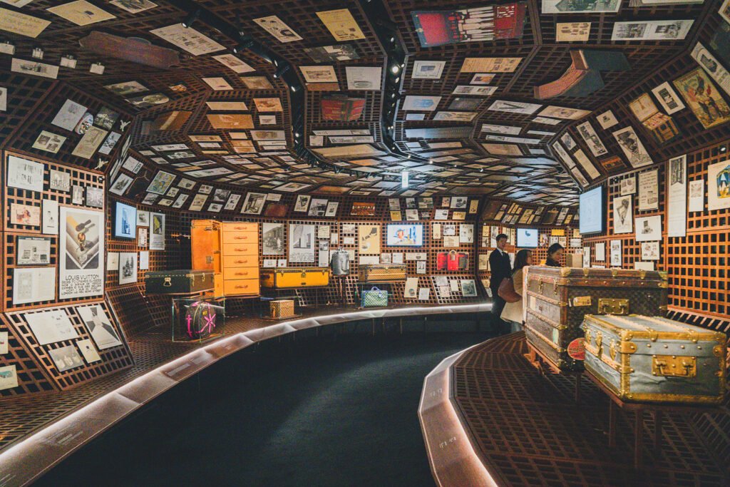 Curved archive room lined ceiling-to-floor with vintage Louis Vuitton ads, catalogs, and antique trunks