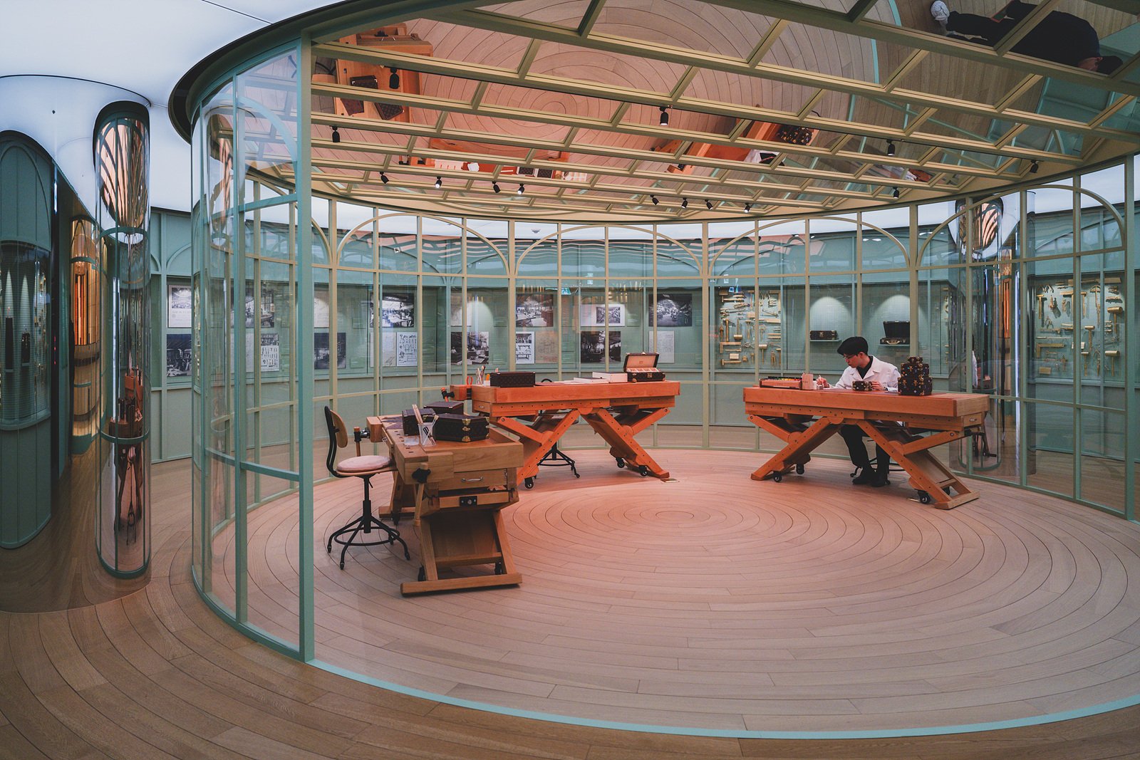 A glass-walled workshop room with wooden workbenches, tools, and a craftsperson working on a trunk