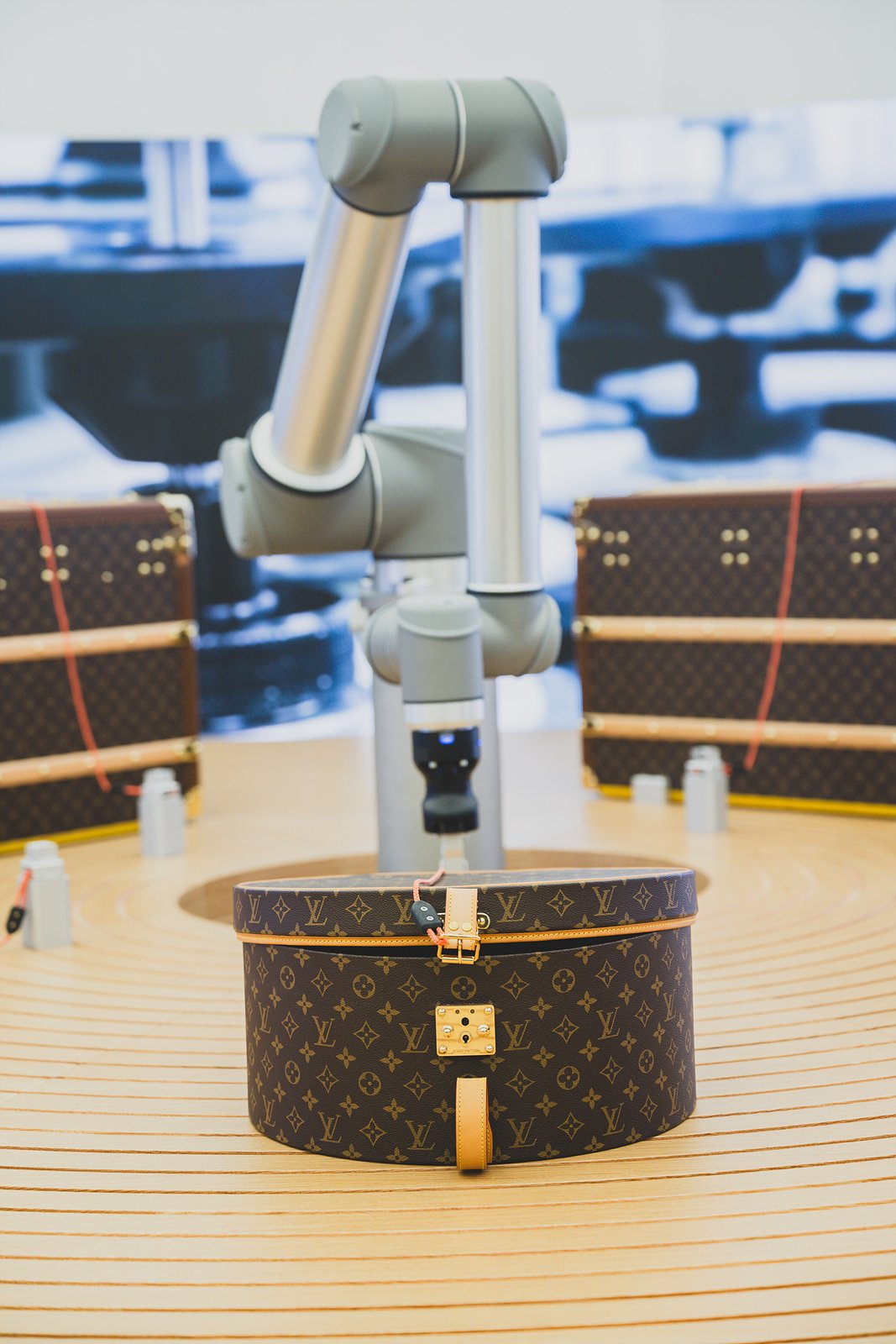 Close-up of the robotic test arm gripping a monogram canvas hatbox-style trunk mid-cycle