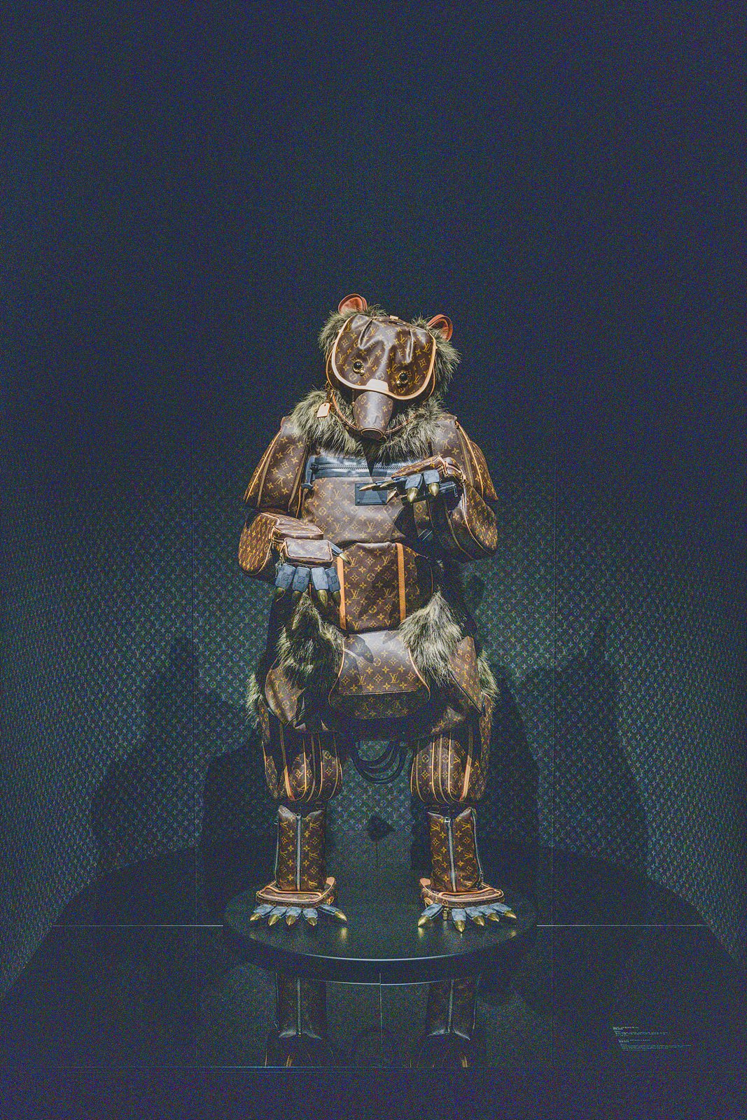 A bear-shaped sculpture built entirely from monogram canvas trunk pieces and fur trim, standing in a dark room
