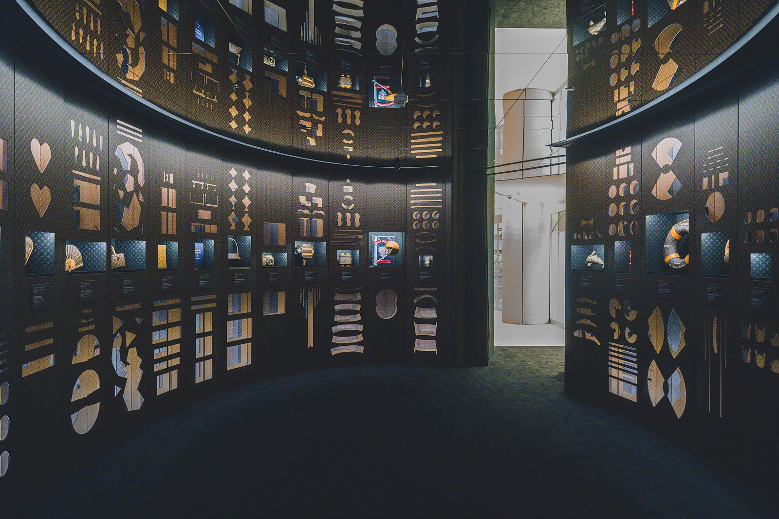 A dimly lit, curved gallery lined with small illuminated display cases showing monogram-pattern objects