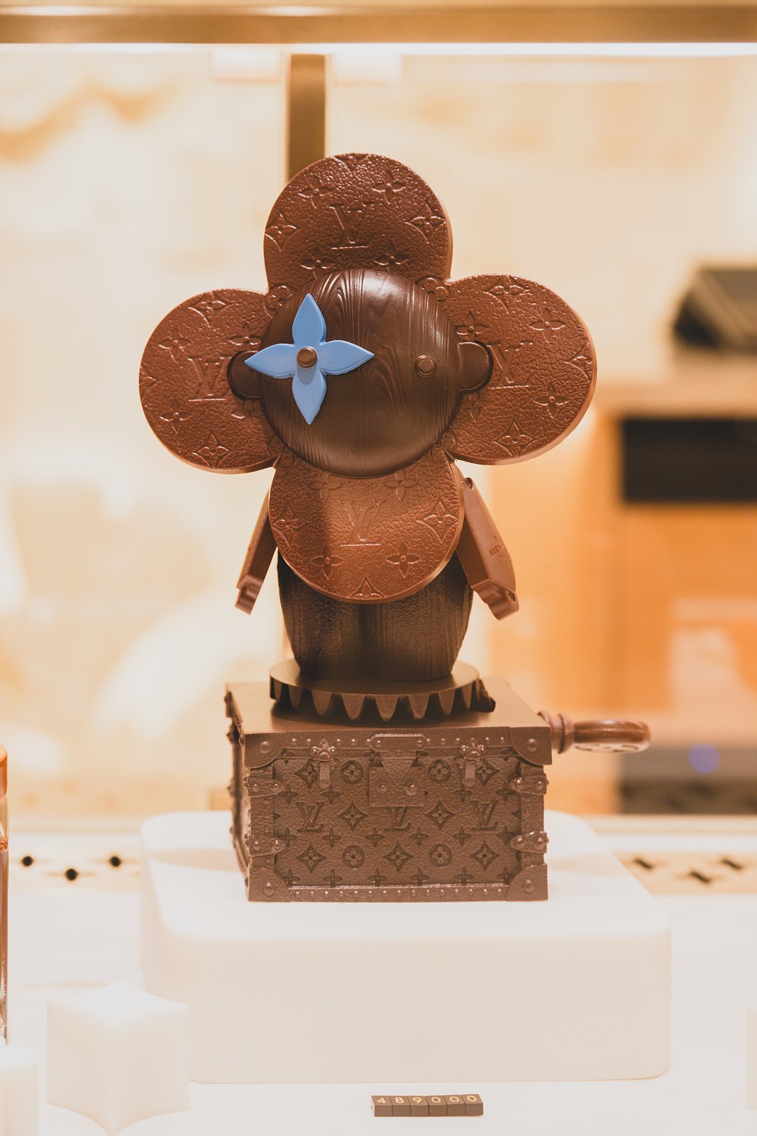 Close-up of a chocolate figure shaped like a four-leaf monogram flower, mounted on a chocolate trunk music box
