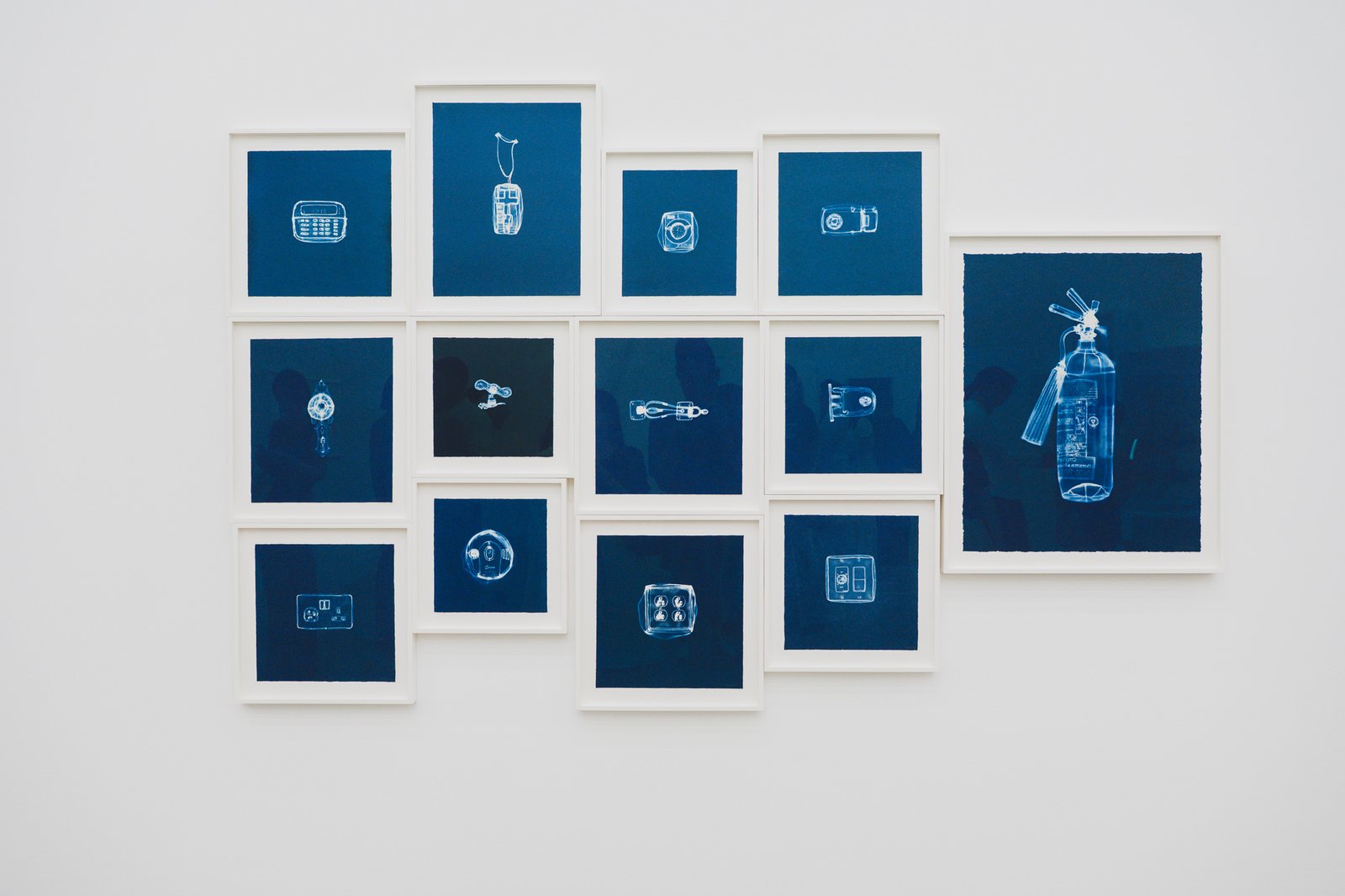 14 blue-toned thread drawings of household objects — fire extinguisher, outlets, doorbell, light switches — hung in a row