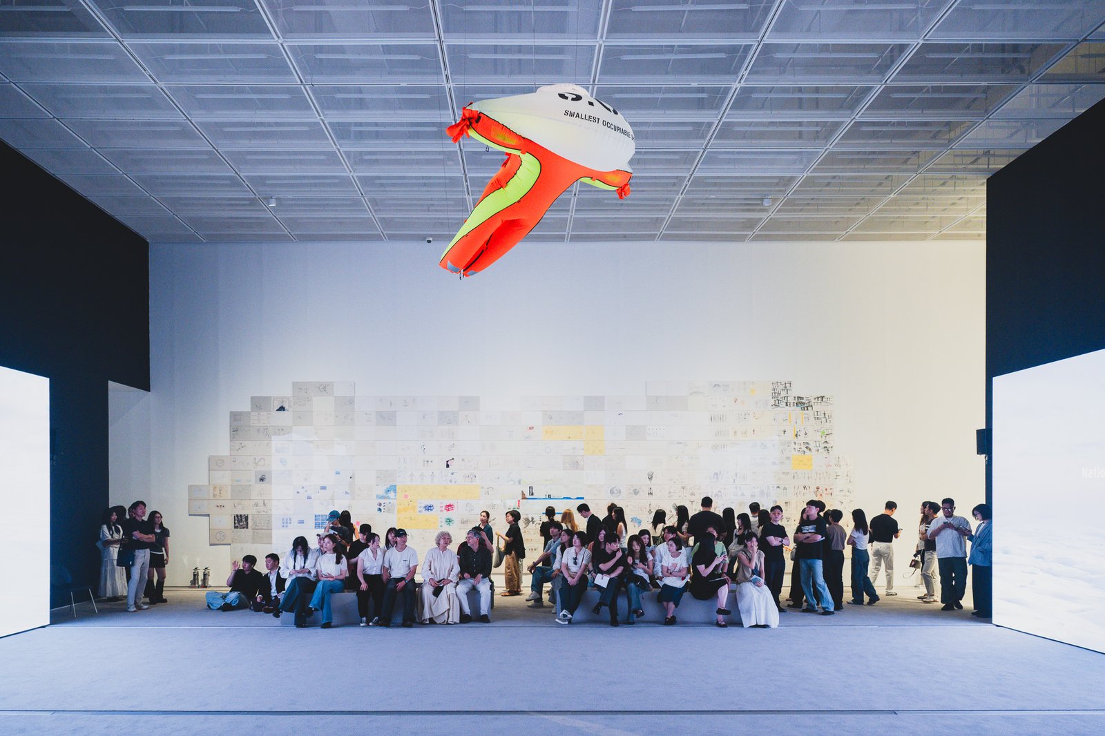Long runway-shaped gallery with orange and green inflatable sculptures overhead, visitors seated on a central bench facing large screens on either wall
