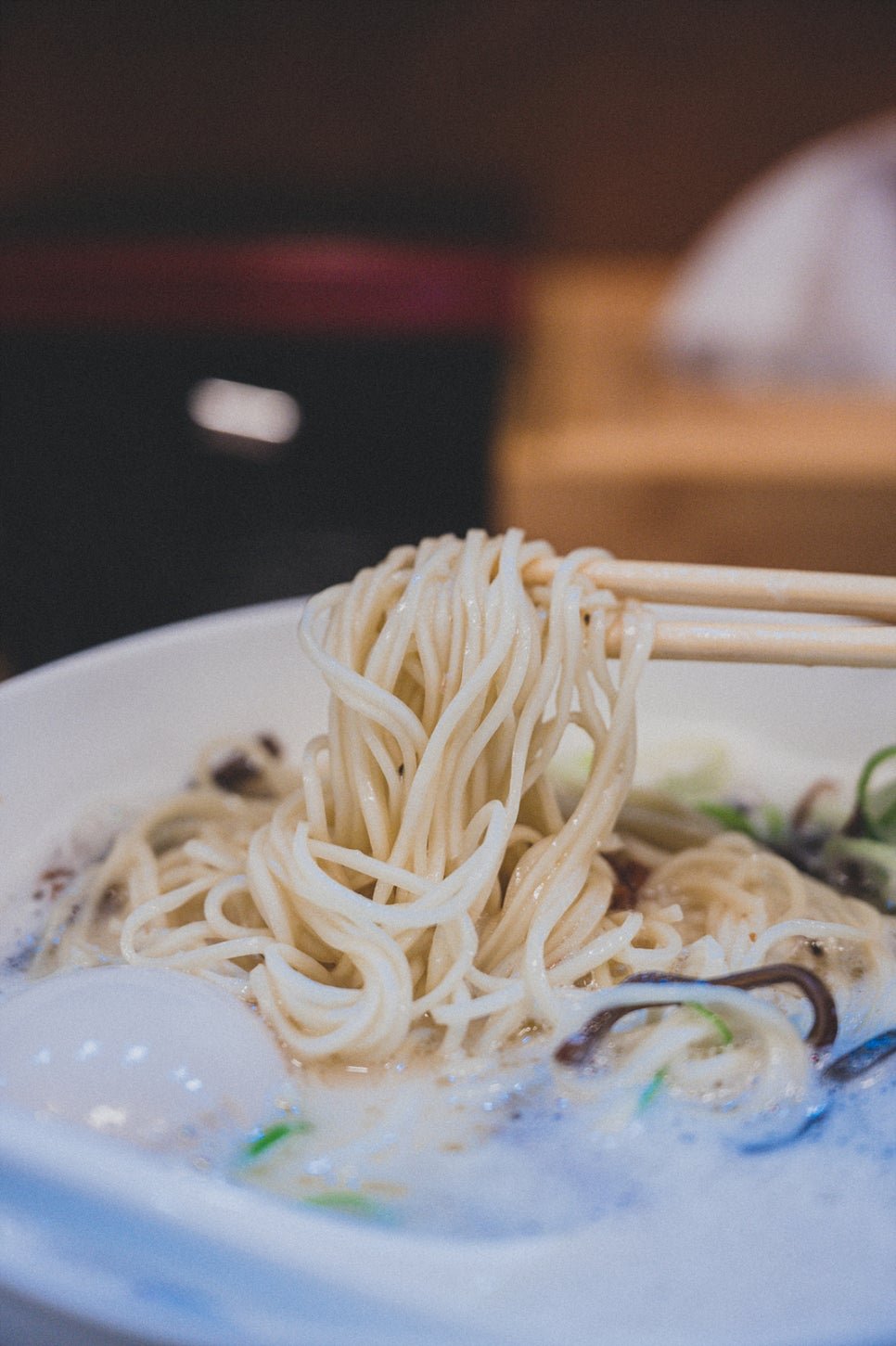 Noodles lifted with chopsticks out of a bowl of pai-tan ramen, showing the texture of the house-made noodles against the creamy broth
