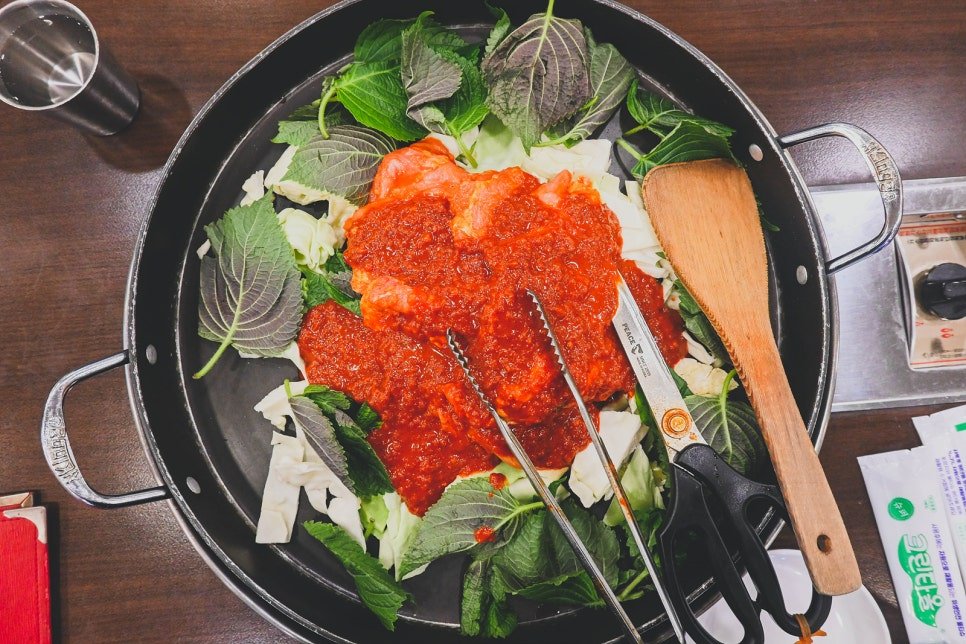 Overhead view of the pan before cooking: red-marinated chicken piled on top of cabbage and perilla leaves, with tongs and a wooden spatula resting on the side