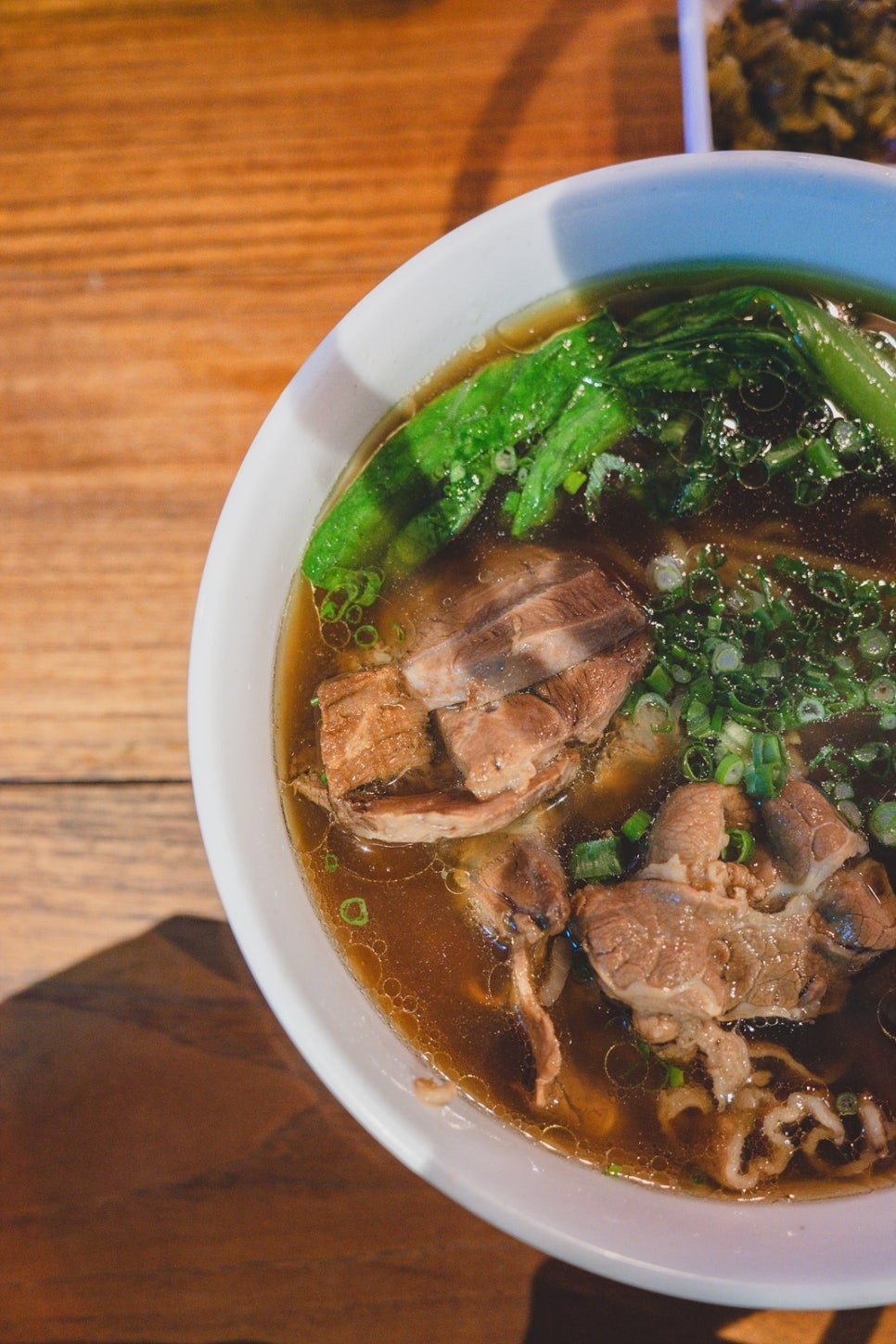 Overhead view of a bowl of beef noodle soup, dark broth with sliced beef, scallions, and greens