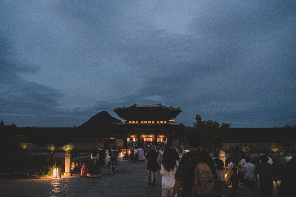 Visitors walking toward Gyeongbokgung's main gate at dusk, the sky still holding orange light behind the palace roofline and Bukhansan mountain in the background