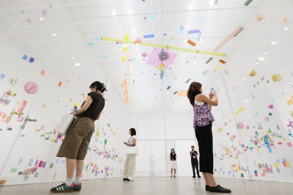Visitors photographing Do Ho Suh's installation at MMCA Seoul, a room with walls covered in colorful hand-drawn rubbings of household hardware — door handles, switches, locks — alongside hanging translucent sculptural objects
