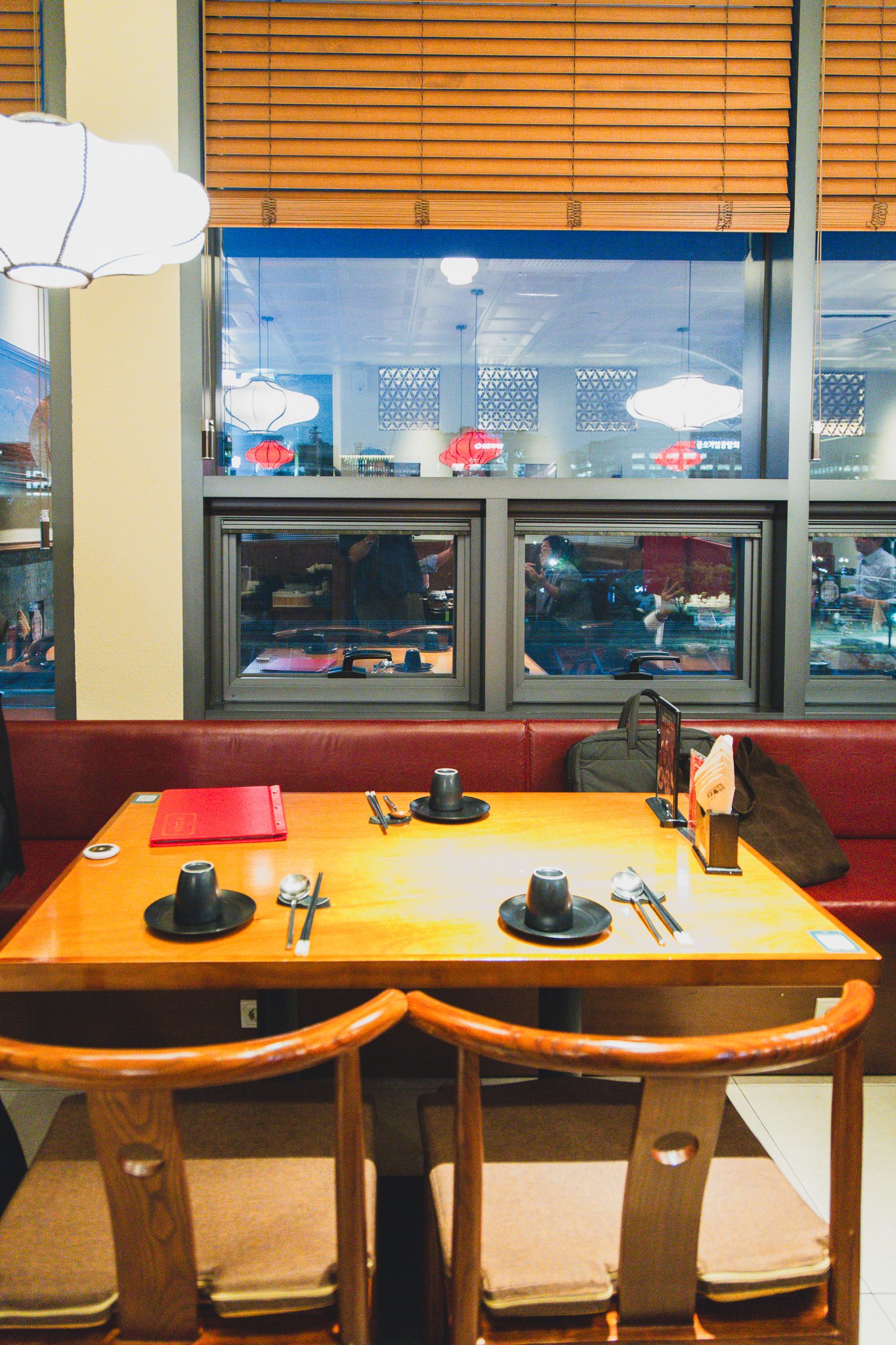 An empty window-side booth table set for two, with a red menu folder, chopsticks, and small side plates, Yeouido's evening skyline visible through the glass behind it