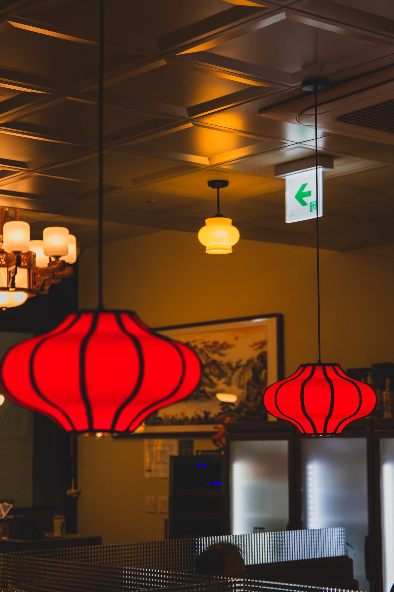 Two red paper lanterns hanging from a patterned ceiling, with a warm-lit pendant lamp and a framed Chinese landscape painting visible in the background