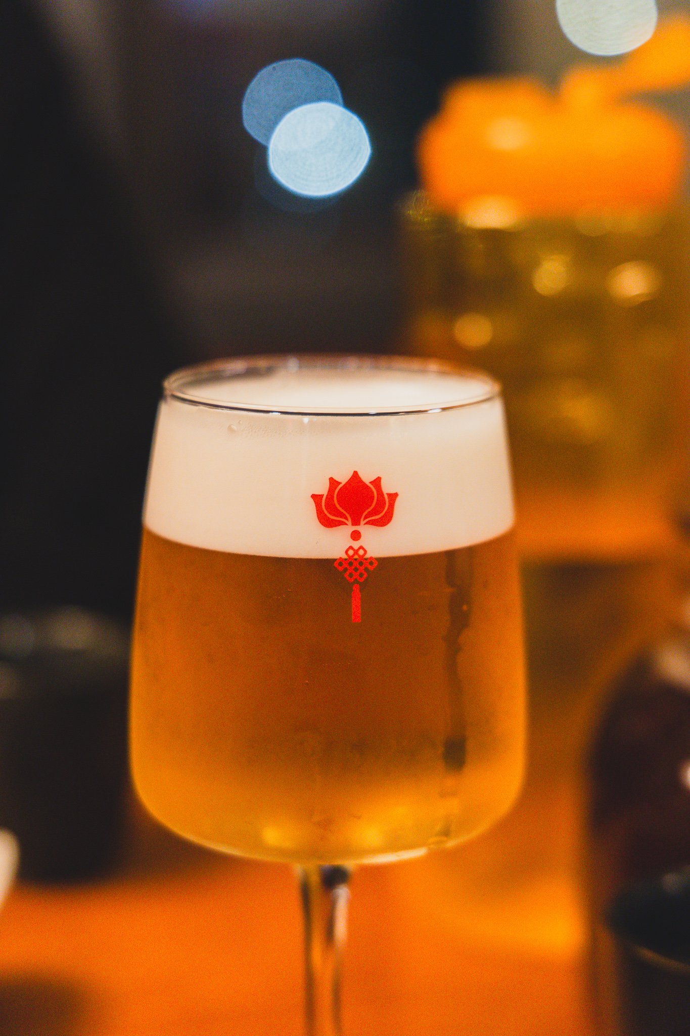 A tall glass of Tsingtao draft beer with a red Rakhian logo printed on the foam line, set against a warmly lit, blurred restaurant background
