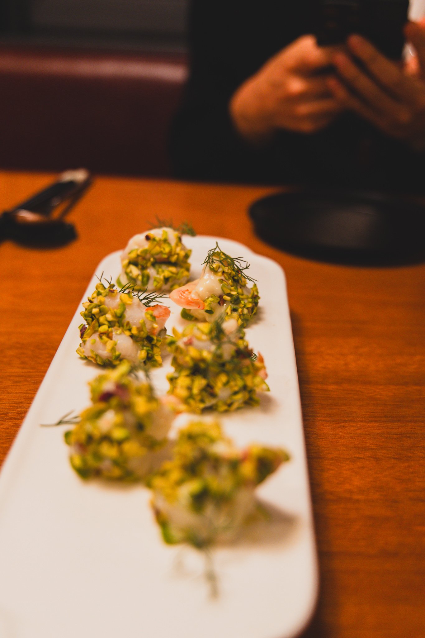 A long white platter with six shrimp fully coated in crushed pistachio, garnished with dill, served on a wooden table