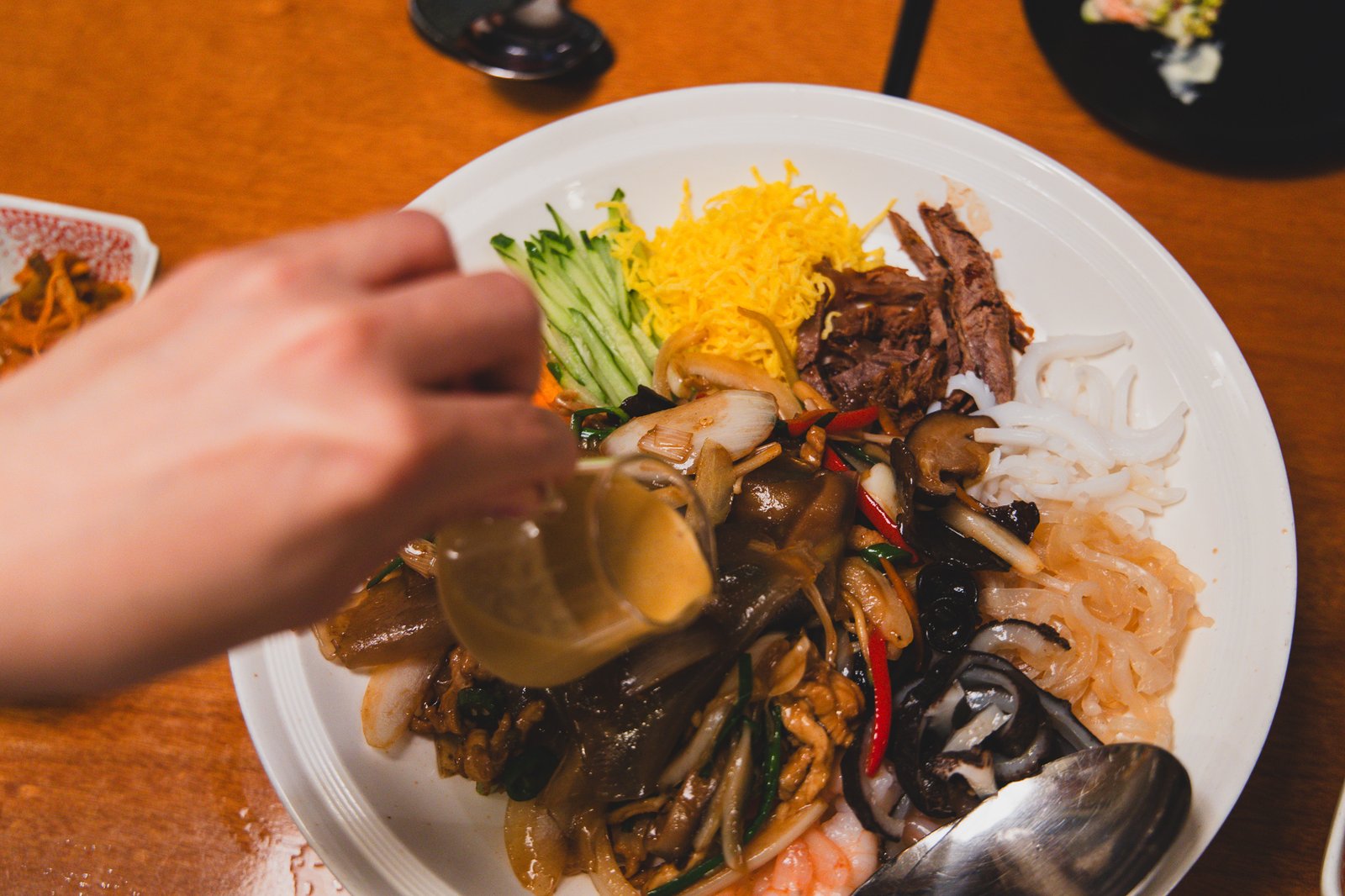A hand pouring mustard sauce from a small glass pitcher over the yangjangpi platter, mixing into the ingredients below