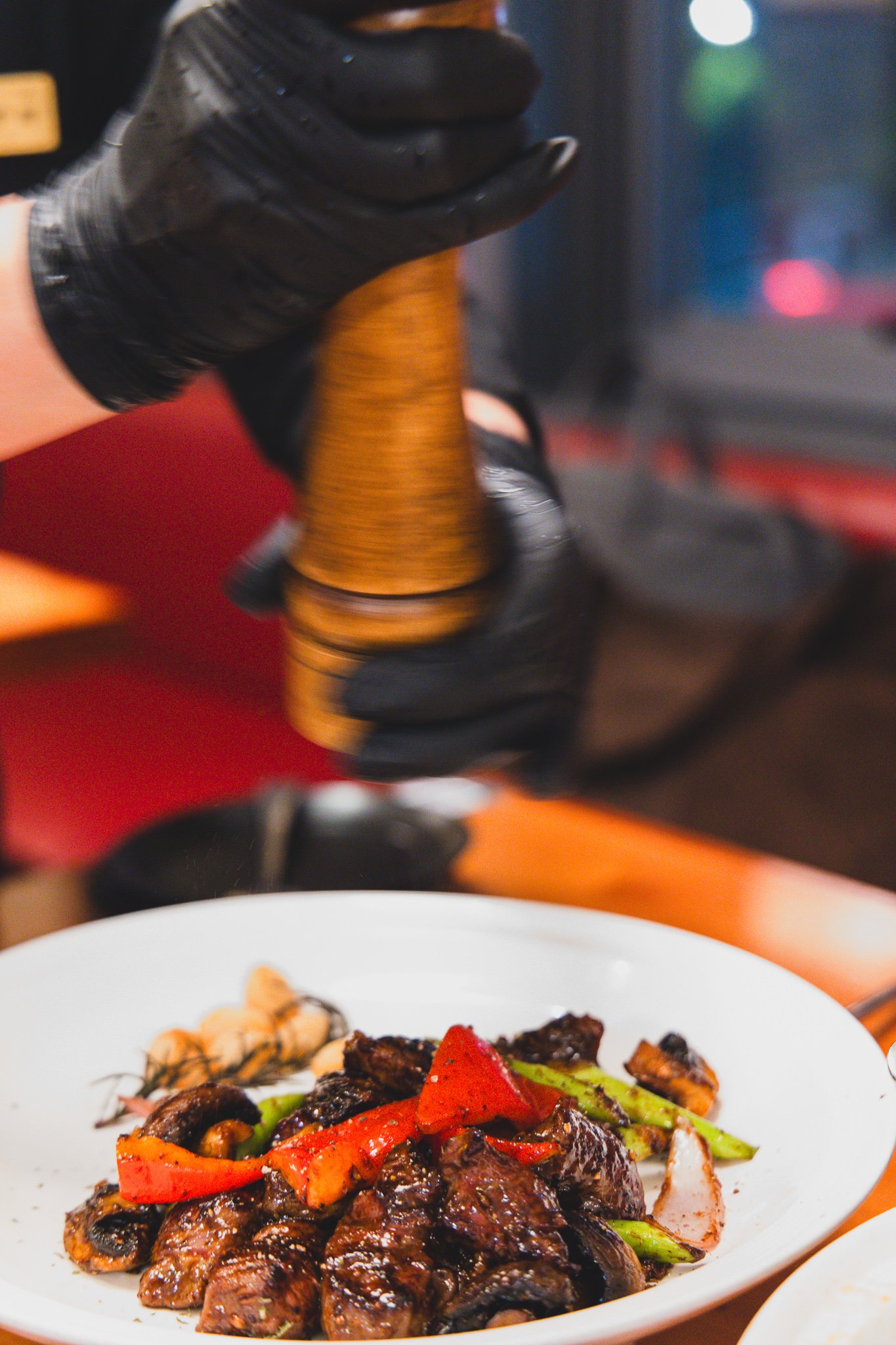A gloved hand grinding a wooden pepper mill directly over a plate of black pepper tenderloin, with red peppers and asparagus visible below