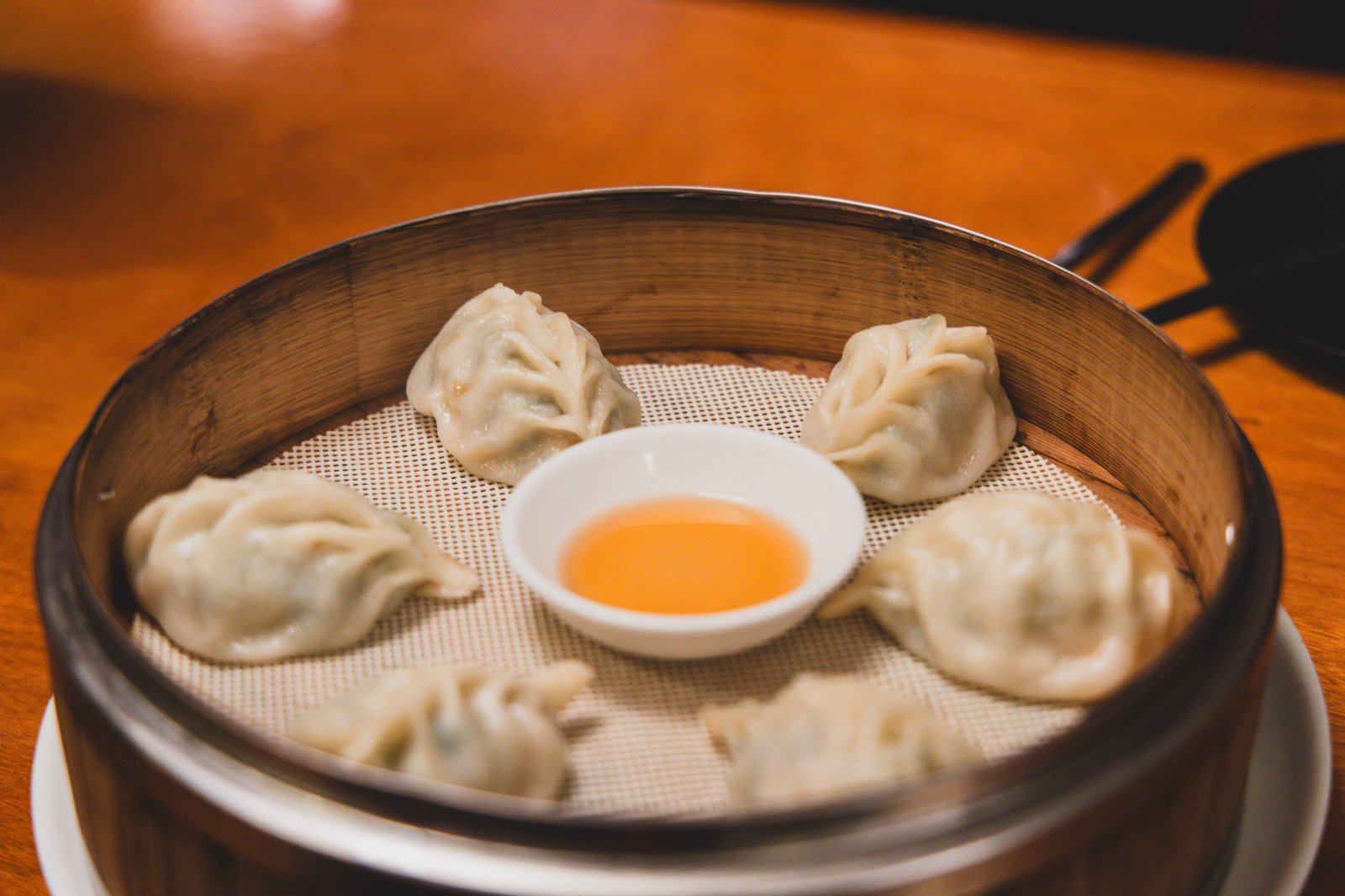 A bamboo steamer holding six pleated shrimp dumplings arranged around a small bowl of amber-colored ginger vinegar dipping sauce