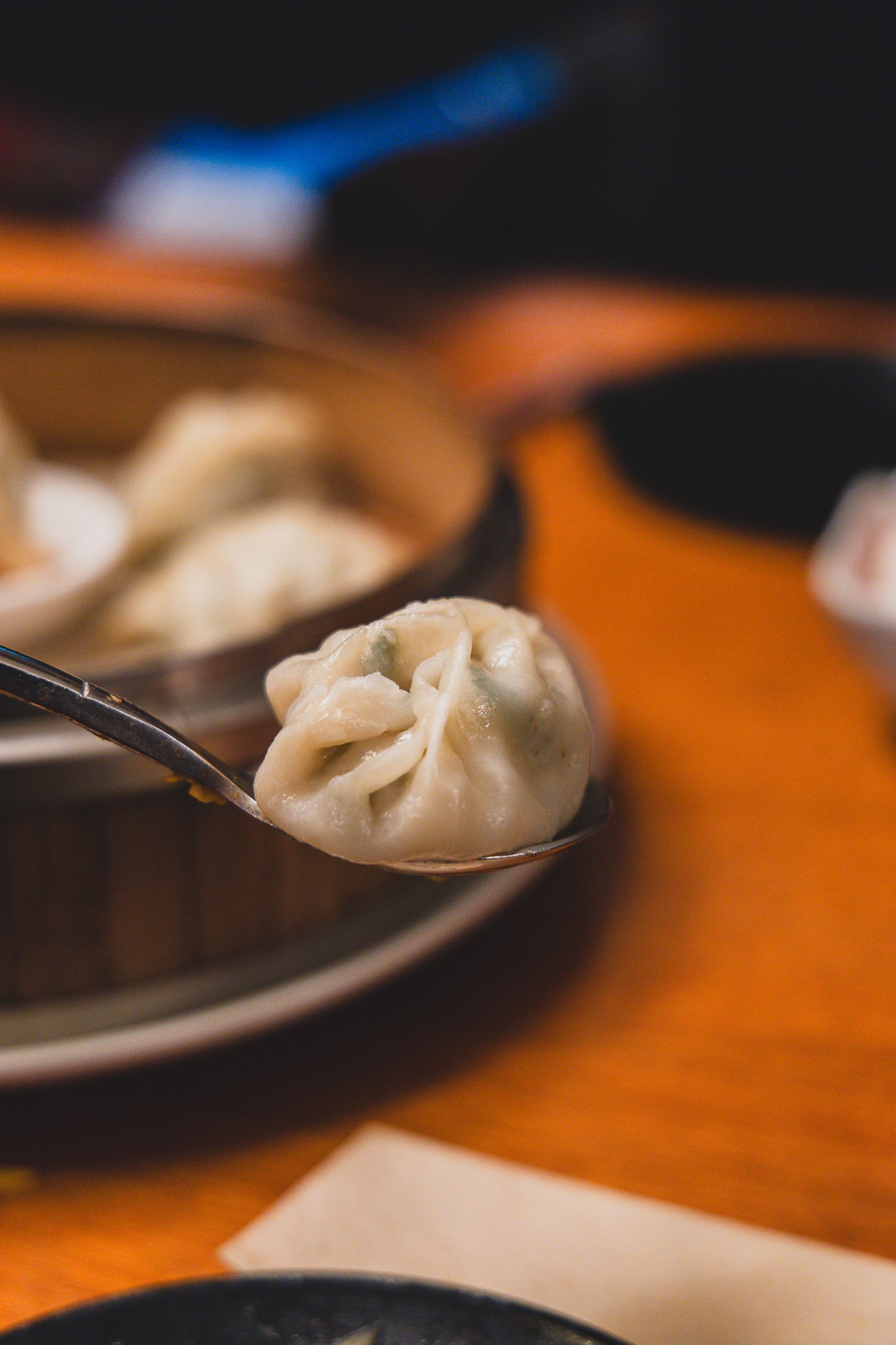 A single dumpling lifted on a spoon, wrapper thin enough to show the shrimp filling inside, steamer and dipping sauce visible in the background