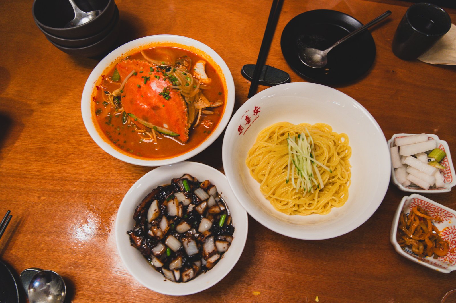 An overhead shot of the full table spread: a whole crab in red spicy broth, a bowl of plain noodles, a dish of black bean sauce with diced pork and onion, and side dishes of pickled radish