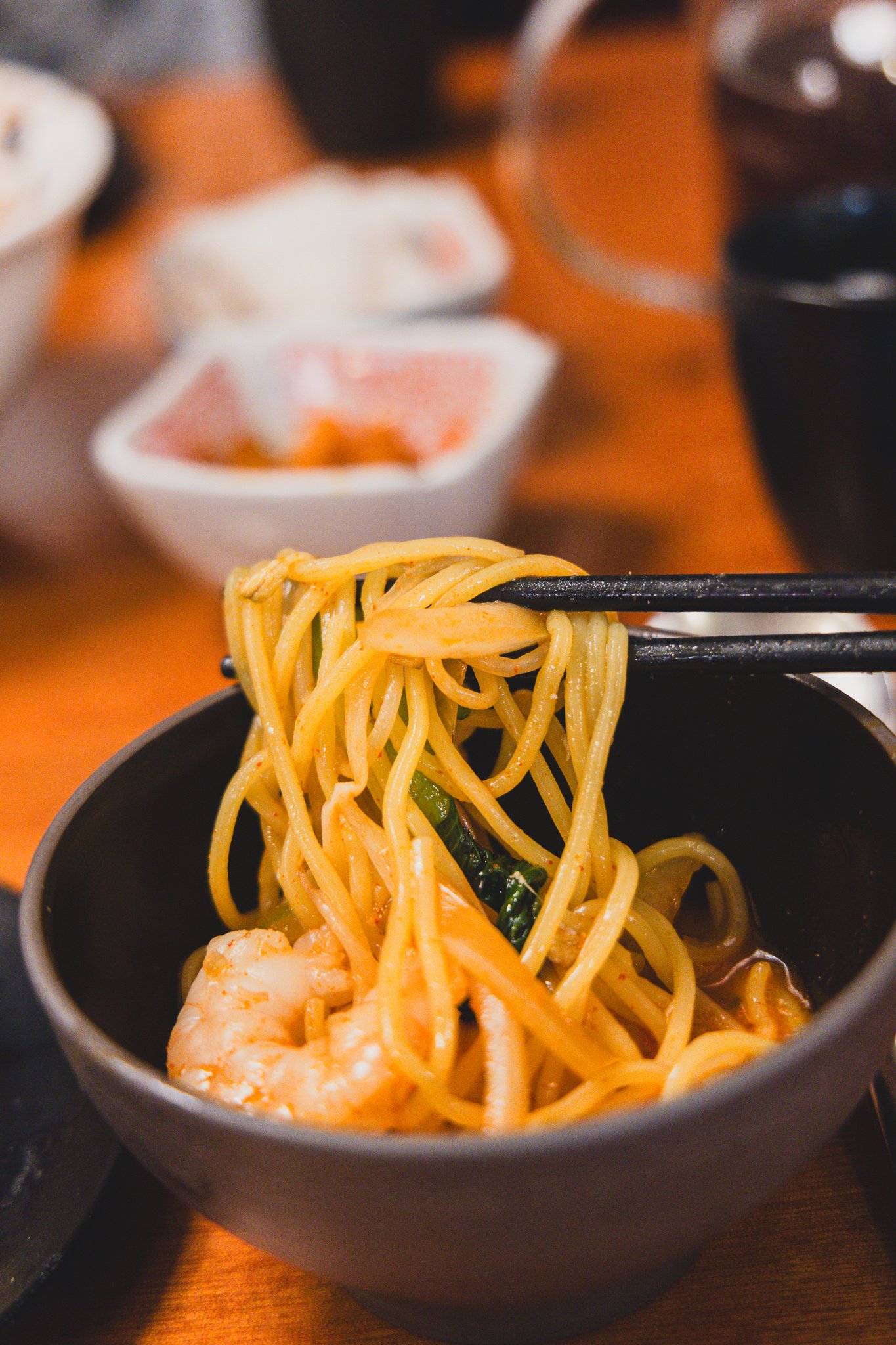 Chopsticks lifting a tangle of noodles out of a dark bowl of spicy jjamppong broth, with a piece of shrimp visible at the bottom