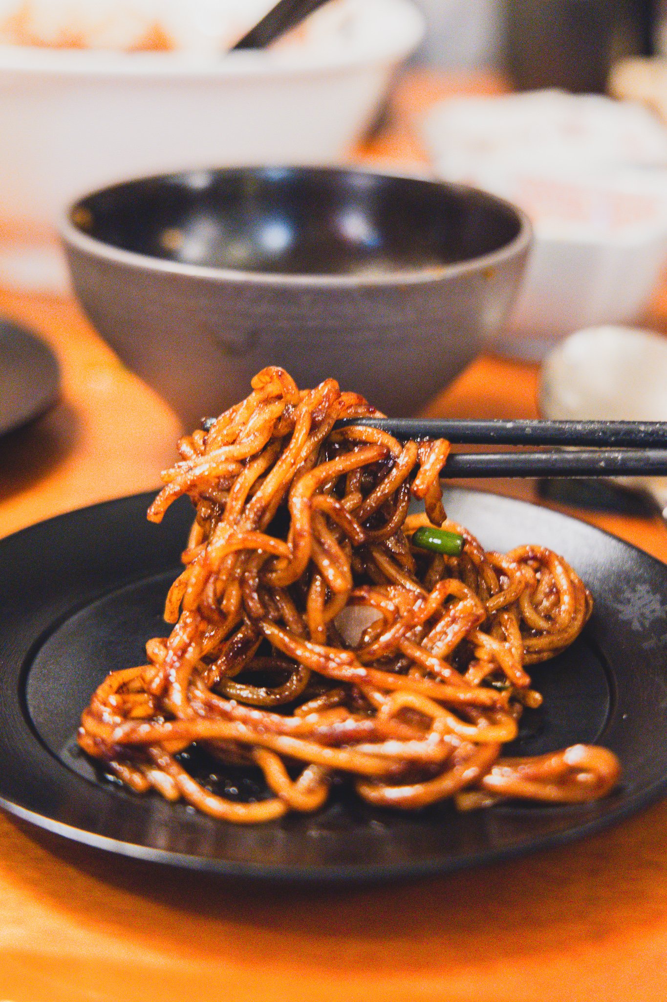 Noodles coated in dark, glossy black bean sauce lifted with chopsticks from a black plate, with visible pieces of pork and scallion