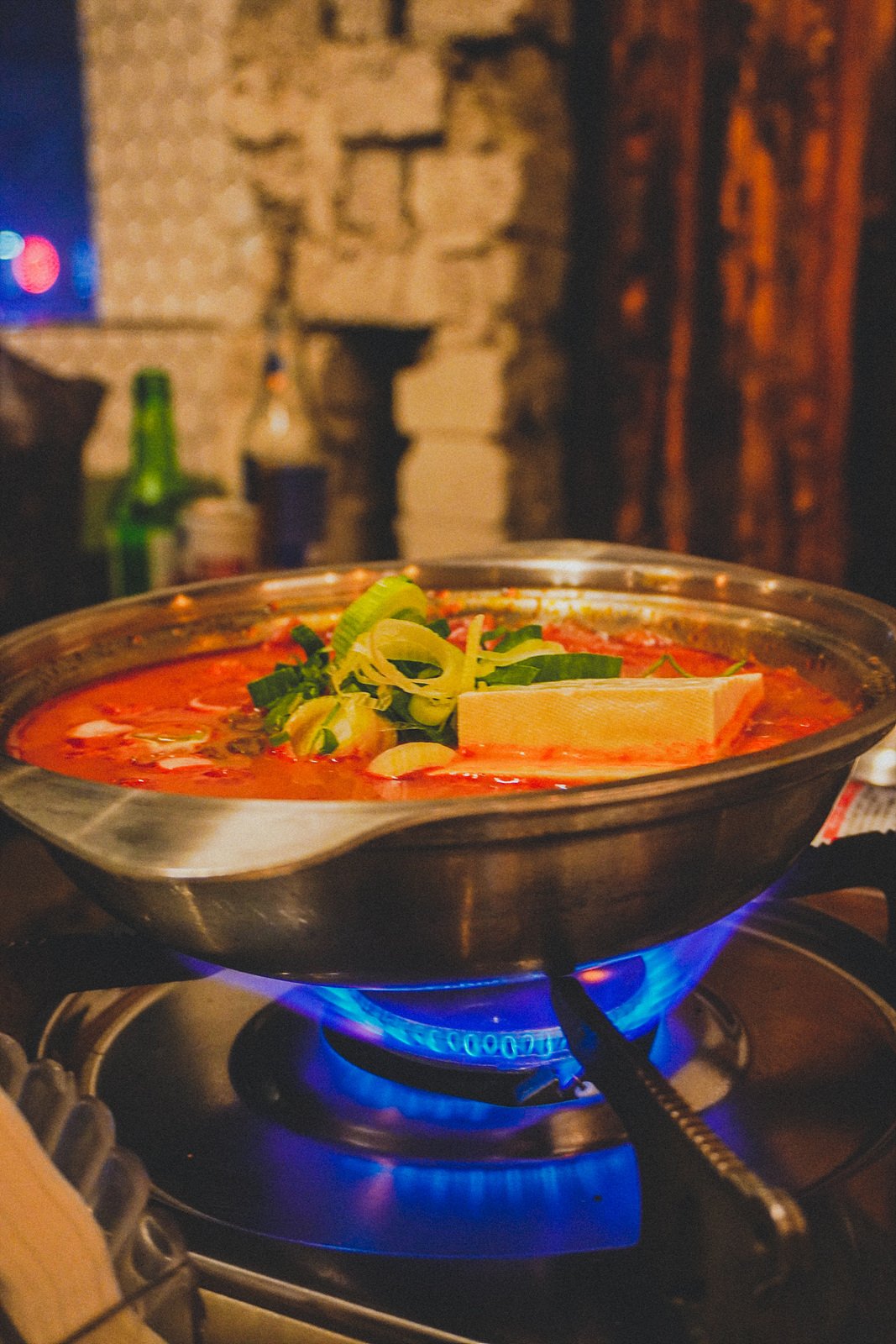 a metal pot of thick red kimchi stew with a block of tofu and scallions, simmering over a blue gas flame