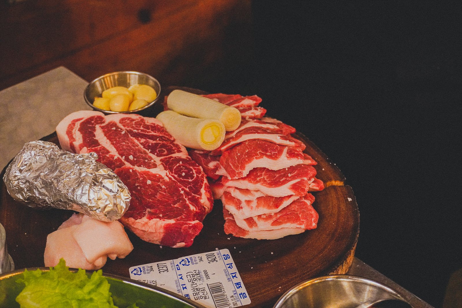 an overhead shot of a wooden serving board with raw cuts of pork: a thick marbled steak-style cut, sliced pork belly, foil-wrapped garlic, and scallions