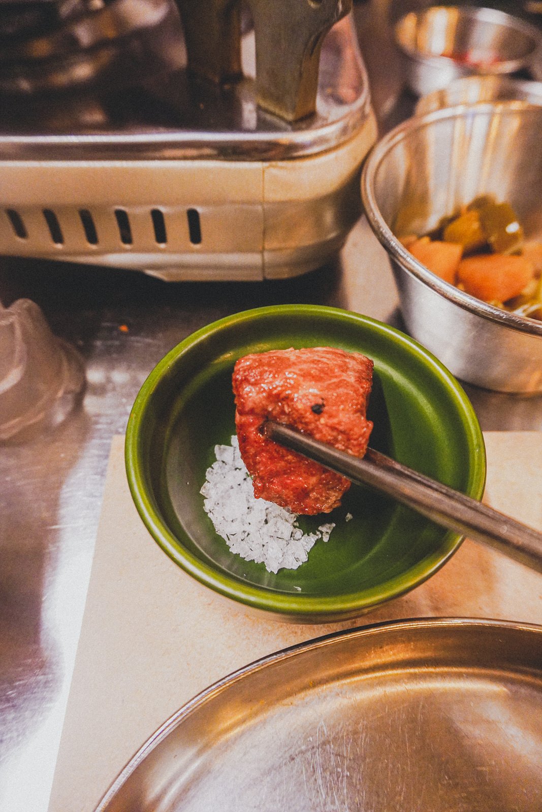 a piece of grilled pork held with chopsticks, dipped into a small pile of coarse salt on a green dish