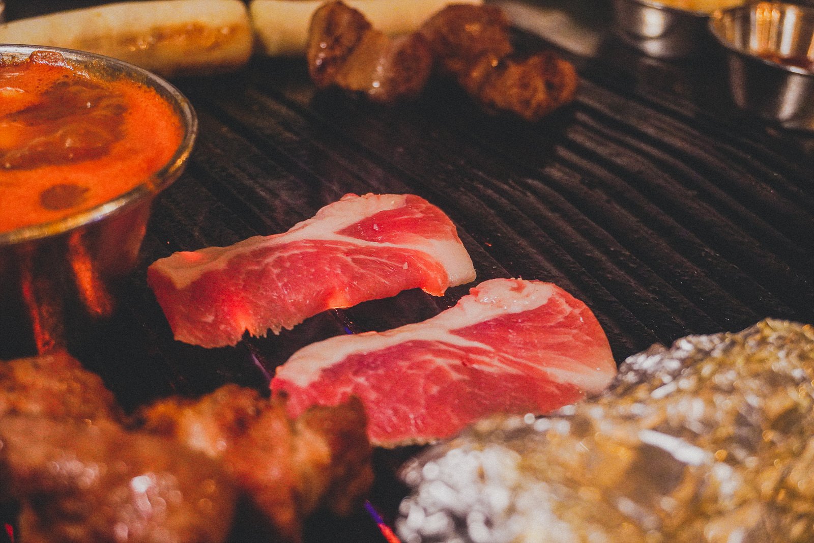 two thick, well-marbled pork slices grilling on a charcoal grate next to golden-browned pieces of pork skin