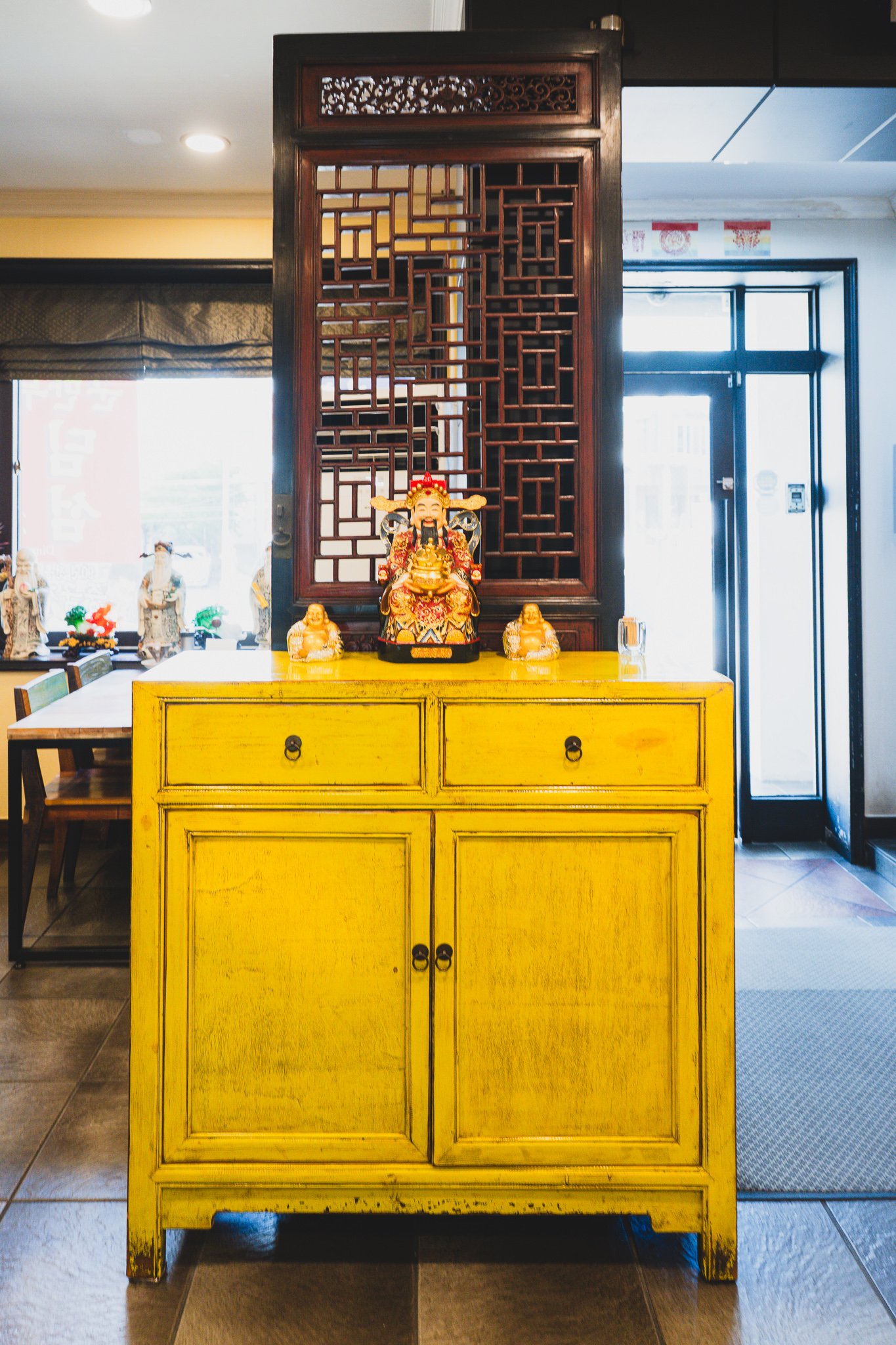 A bright yellow lacquered cabinet topped with a wealth god statue and two smiling Buddha figures, set against a carved wooden screen