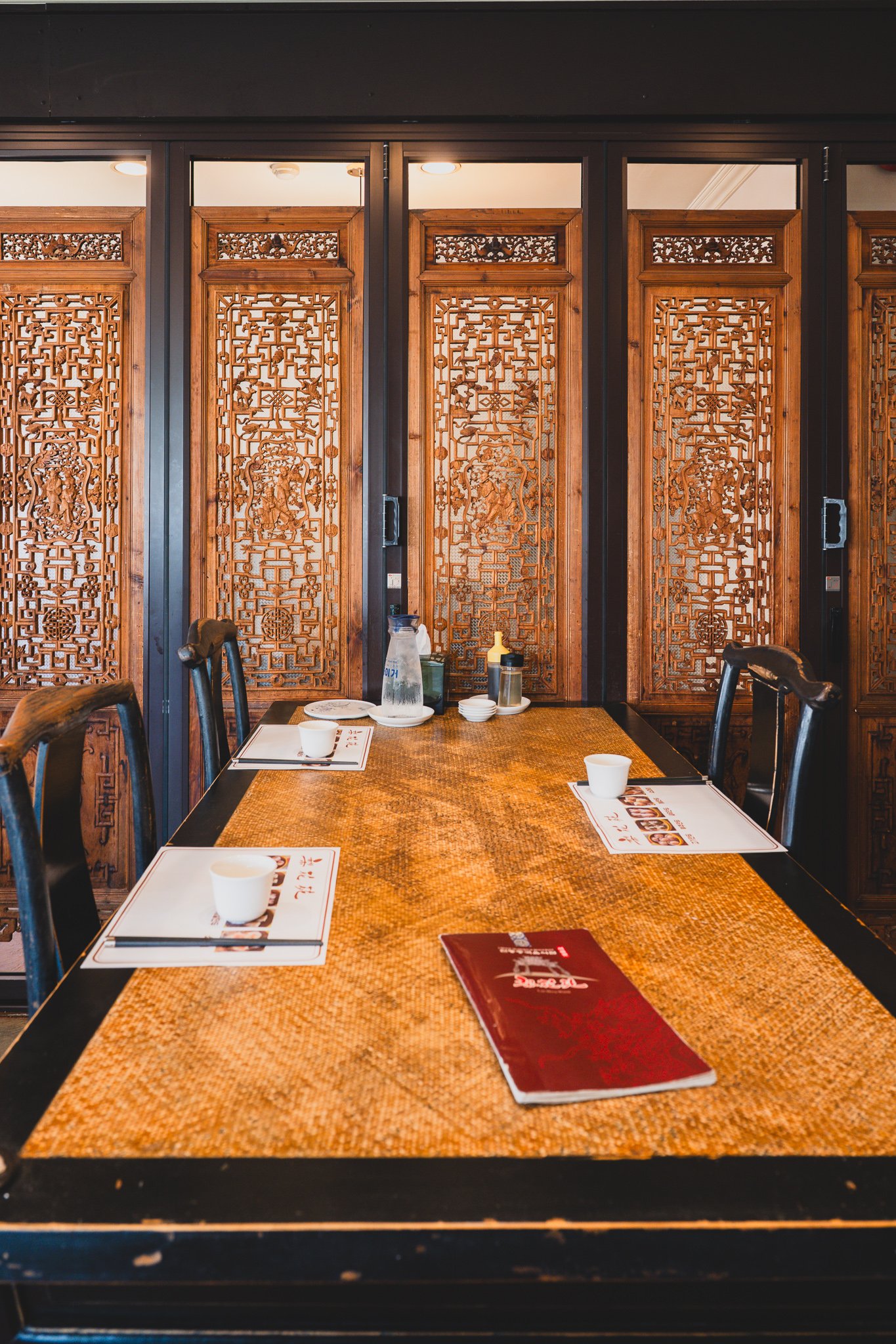 A private dining room table set with placemats, teacups, and a red menu booklet, enclosed by ornate carved wooden screen panels