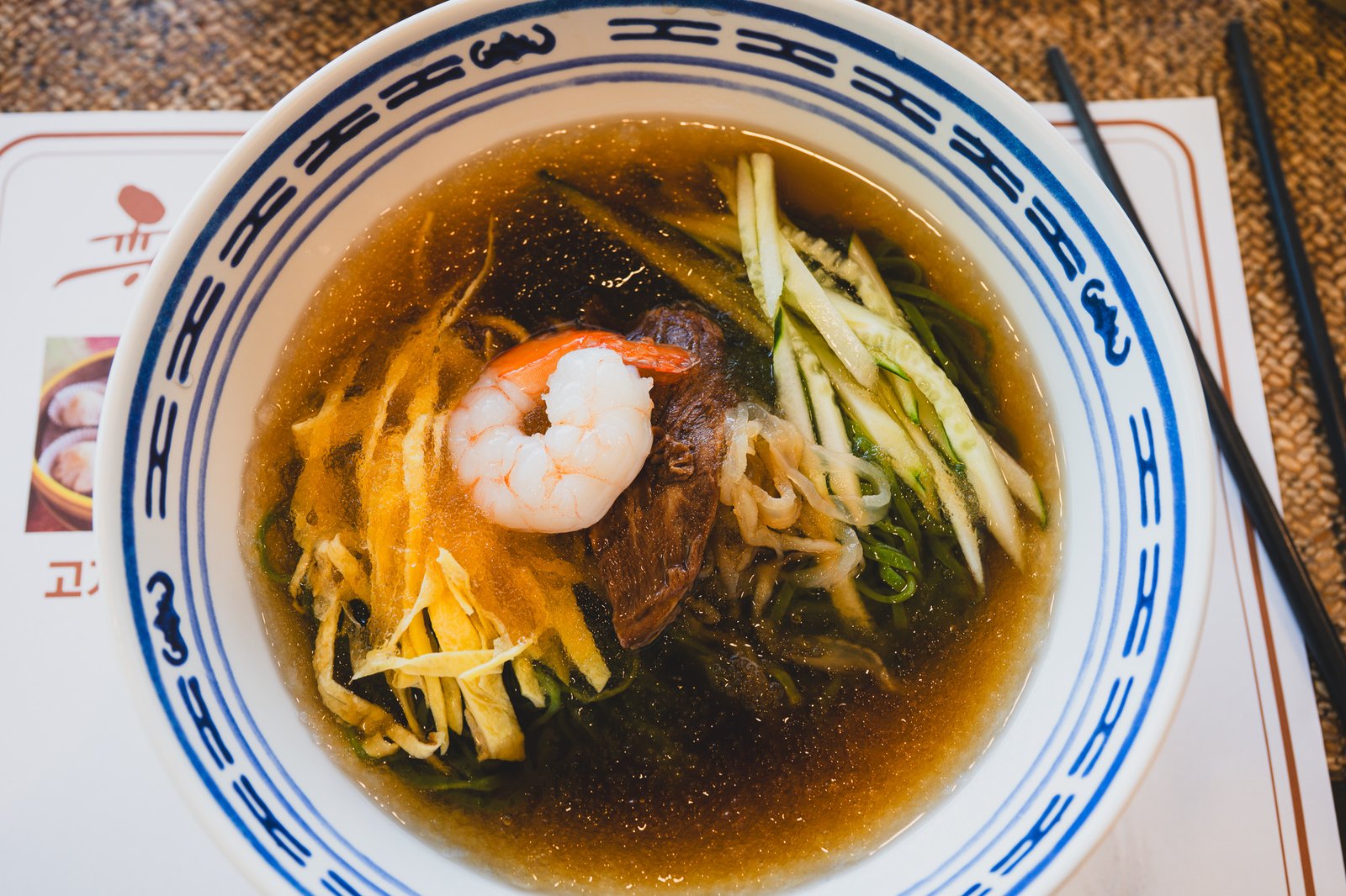 Bowl of jade-green Ihwawon cold noodles topped with shrimp, sliced beef, julienned cucumber, and shredded egg, in a savory brown broth
