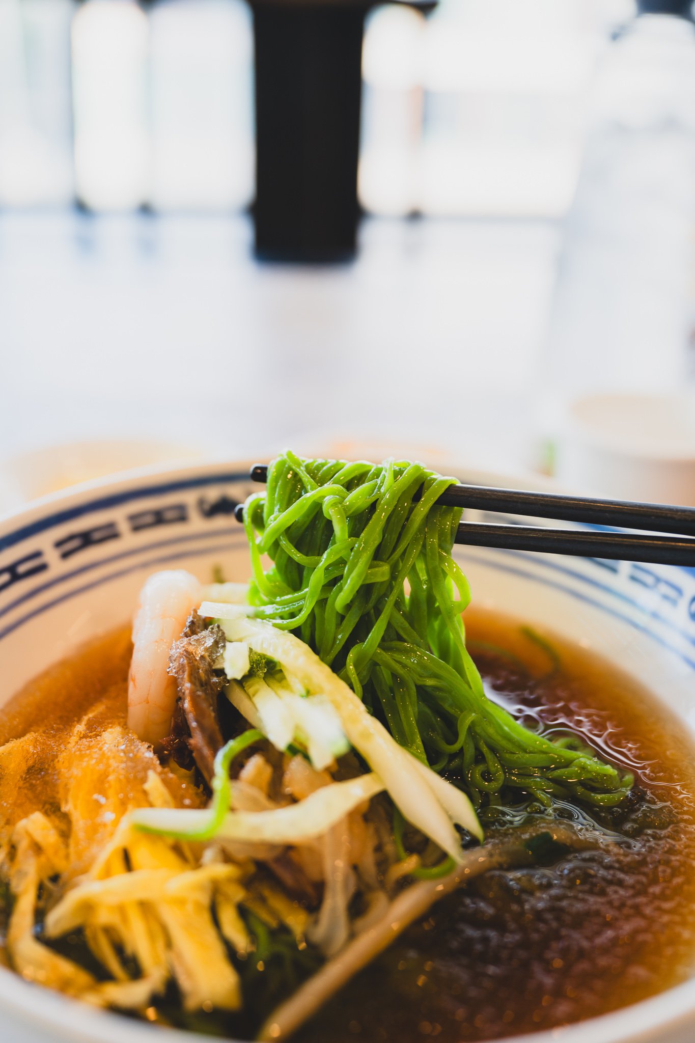 Close-up of chopsticks lifting a tangle of bright green jade noodles out of the bowl, broth dripping back down