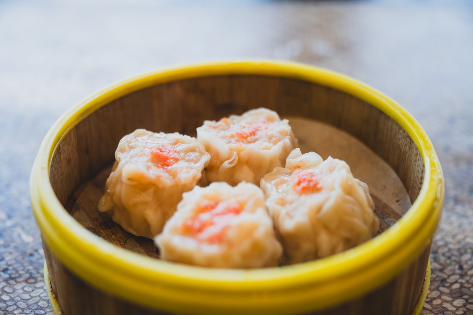 Four shumai dumplings topped with orange roe, served in a yellow-rimmed bamboo steamer basket
