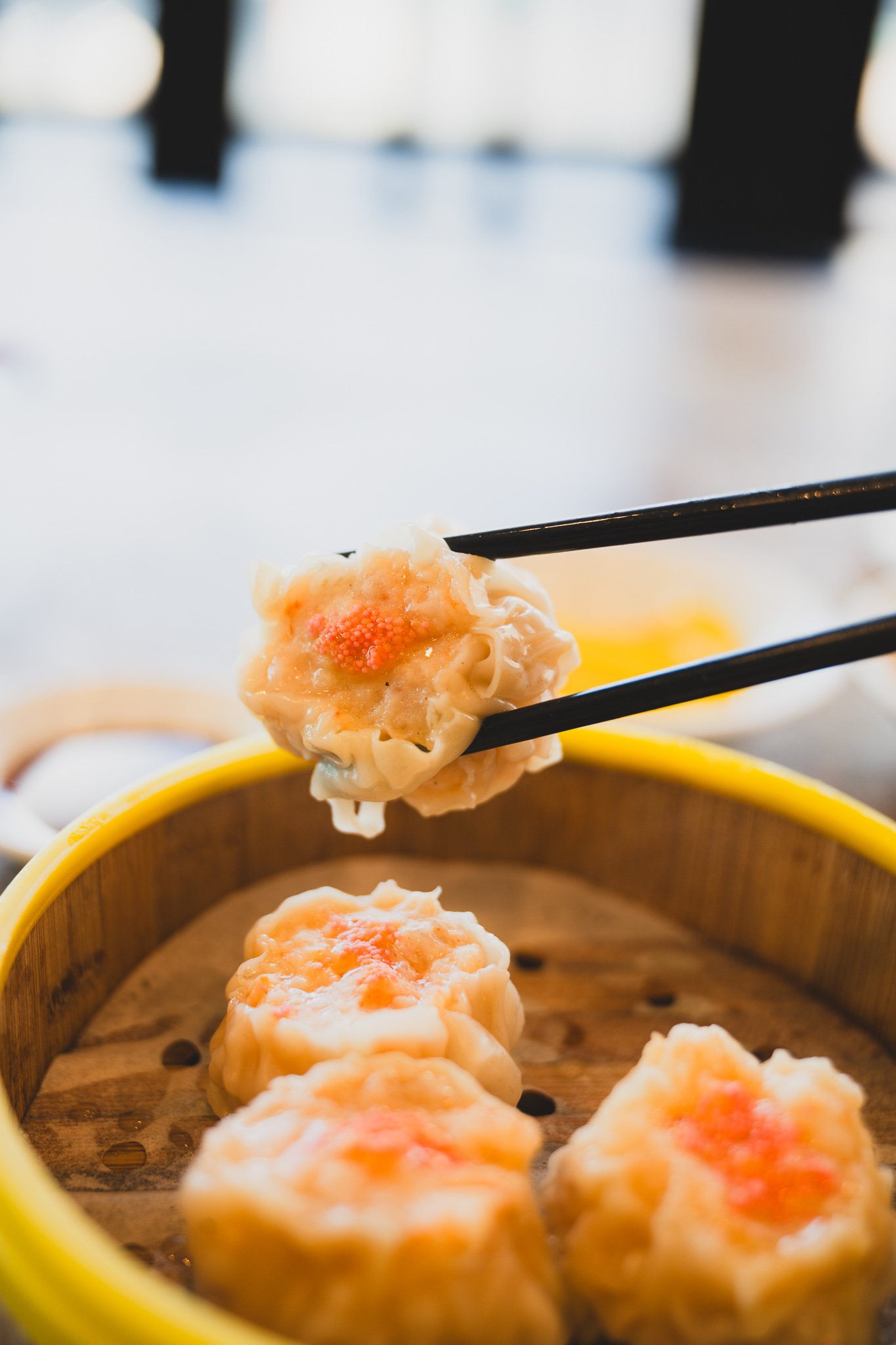 Chopsticks lifting a single shumai dumpling toward the camera, more dumplings visible in the steamer basket below