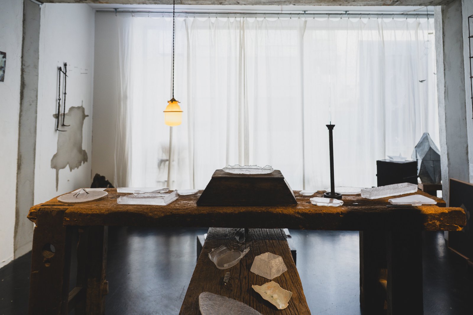 A weathered wooden workbench inside OJA CRAFT displaying several translucent cast-glass plates and objects, lit by a hanging bulb against sheer curtains