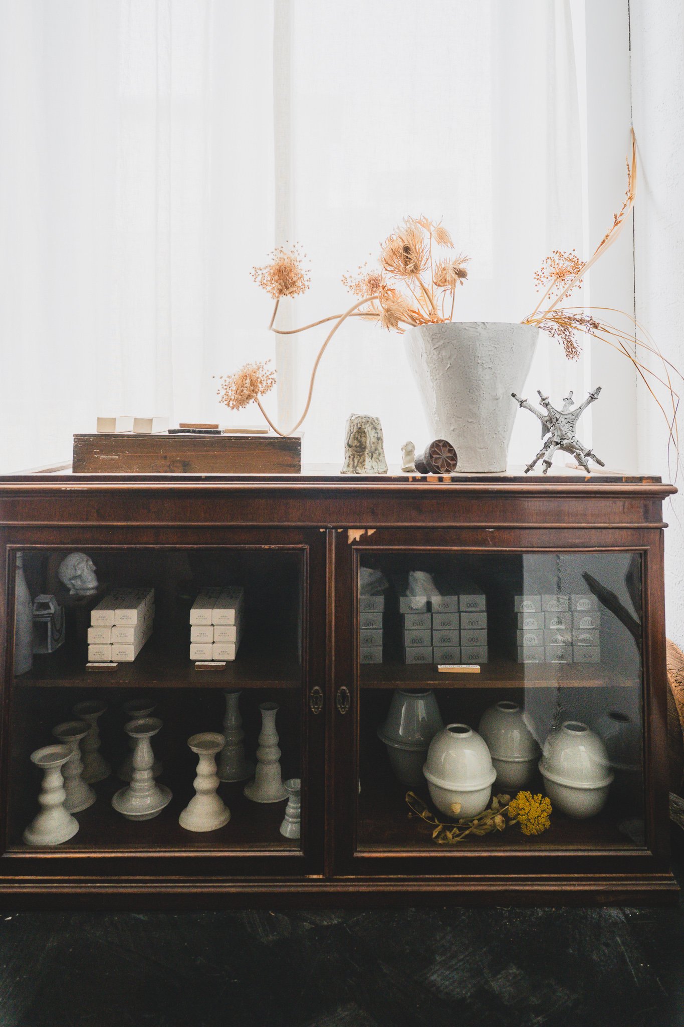 Antique glass-fronted wooden cabinet displaying ceramic candlesticks and vases, topped with dried flower arrangements and small glass and ceramic objects