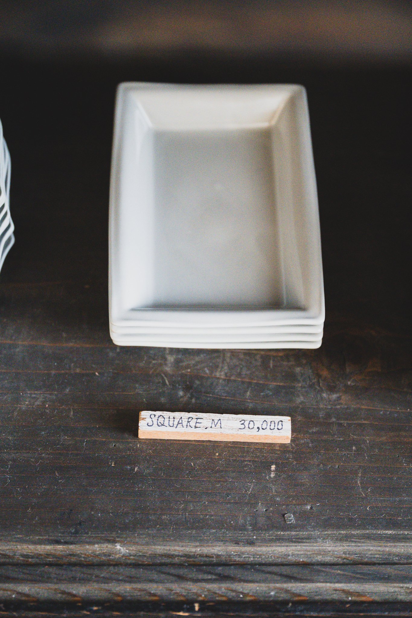 A stack of square white ceramic plates on a dark wooden shelf, with a handwritten wooden price tag reading "Square M 30,000"
