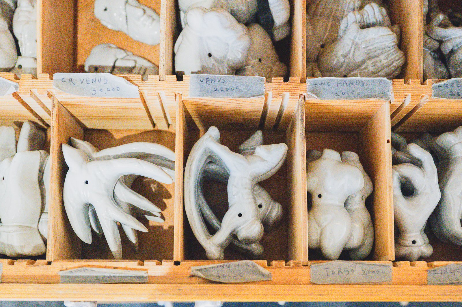 Wooden drawer unit filled with body-shaped ceramic mold parts, each cubby hand-labeled with names like "Venus," "Two Hands," and "Torso" and handwritten prices