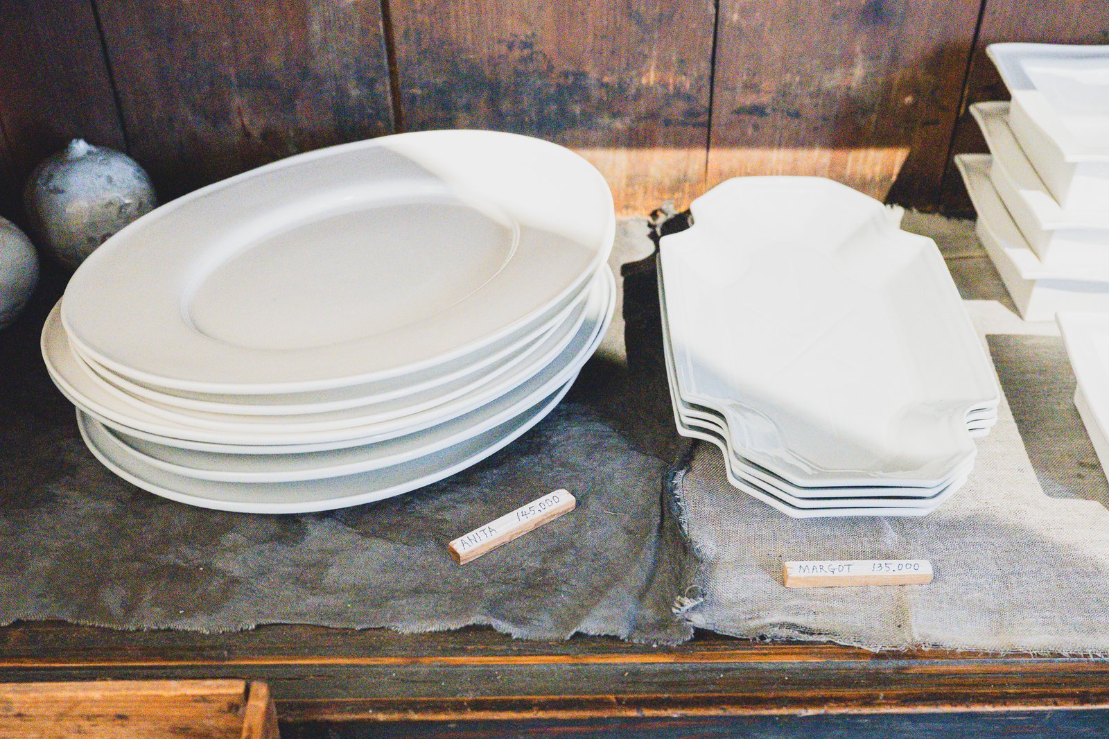 Stacked white ceramic oval platters and scalloped platters on an antique wooden shelf, each with a handwritten price tag reading "Anita 145,000" and "Margot 135,000"